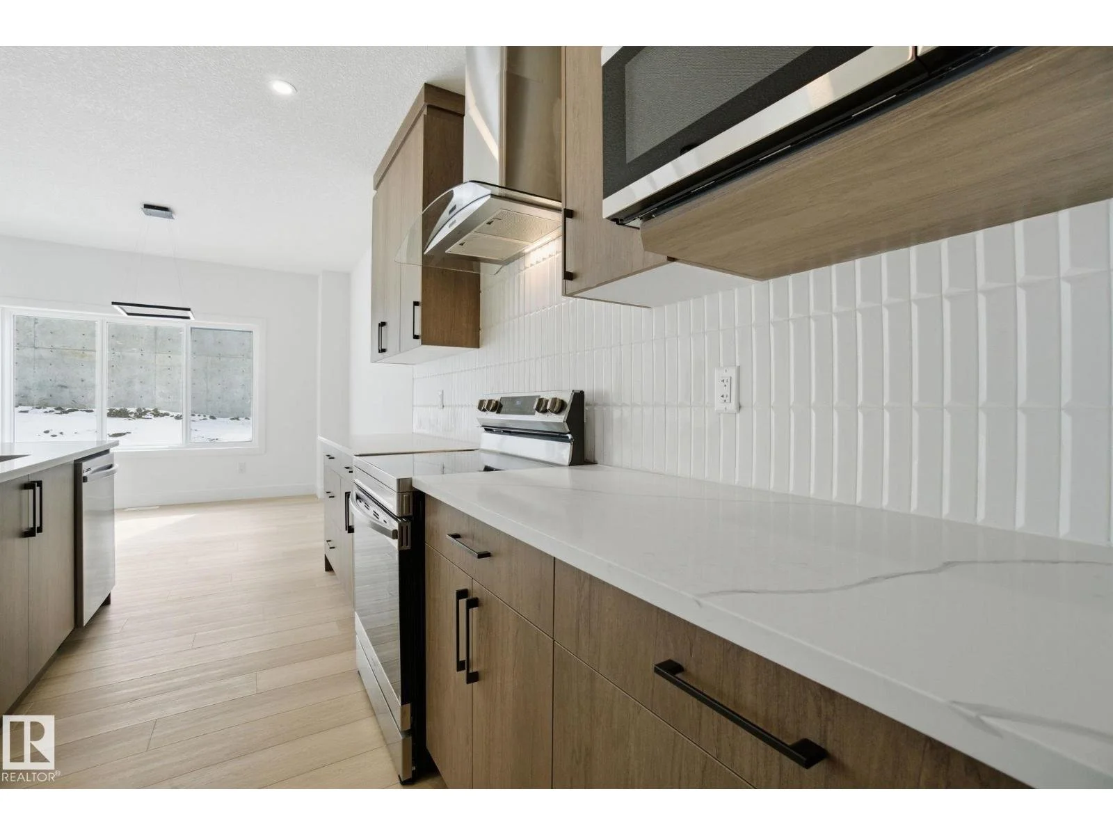 Modern kitchen with wood cabinetry, white countertops, white tiled backsplash, stainless steel appliances, light wood flooring, and a large window letting in natural light.