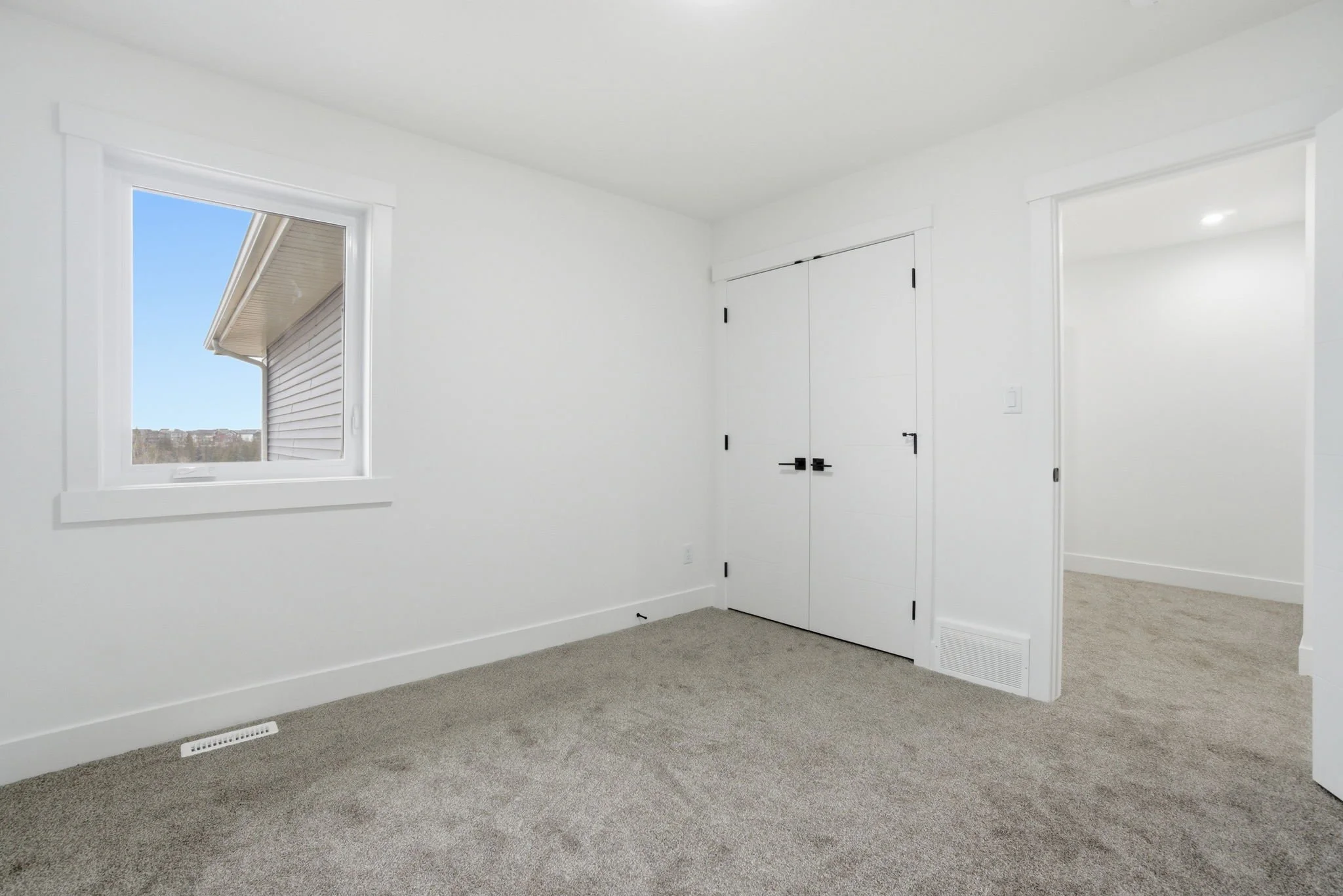 Empty white room with a window showing a view of a house and blue sky, and a closed closet door, beige carpet, and a floor vent.