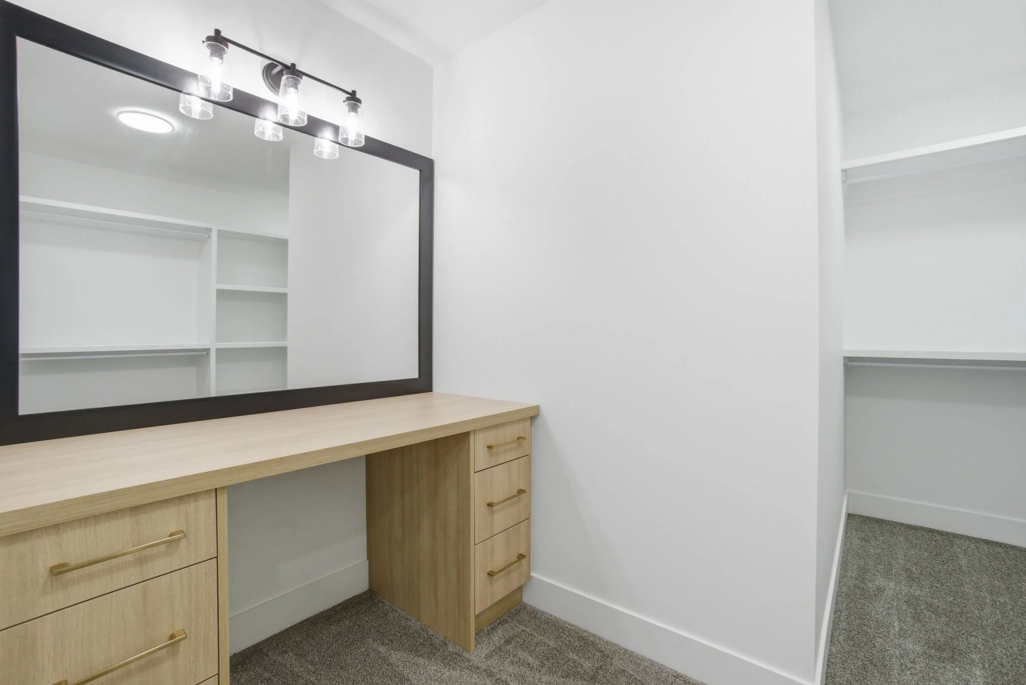 A modern empty walk-in closet with a large mirror, a light fixture with multiple bulbs, a light wooden countertop, and white shelving units.