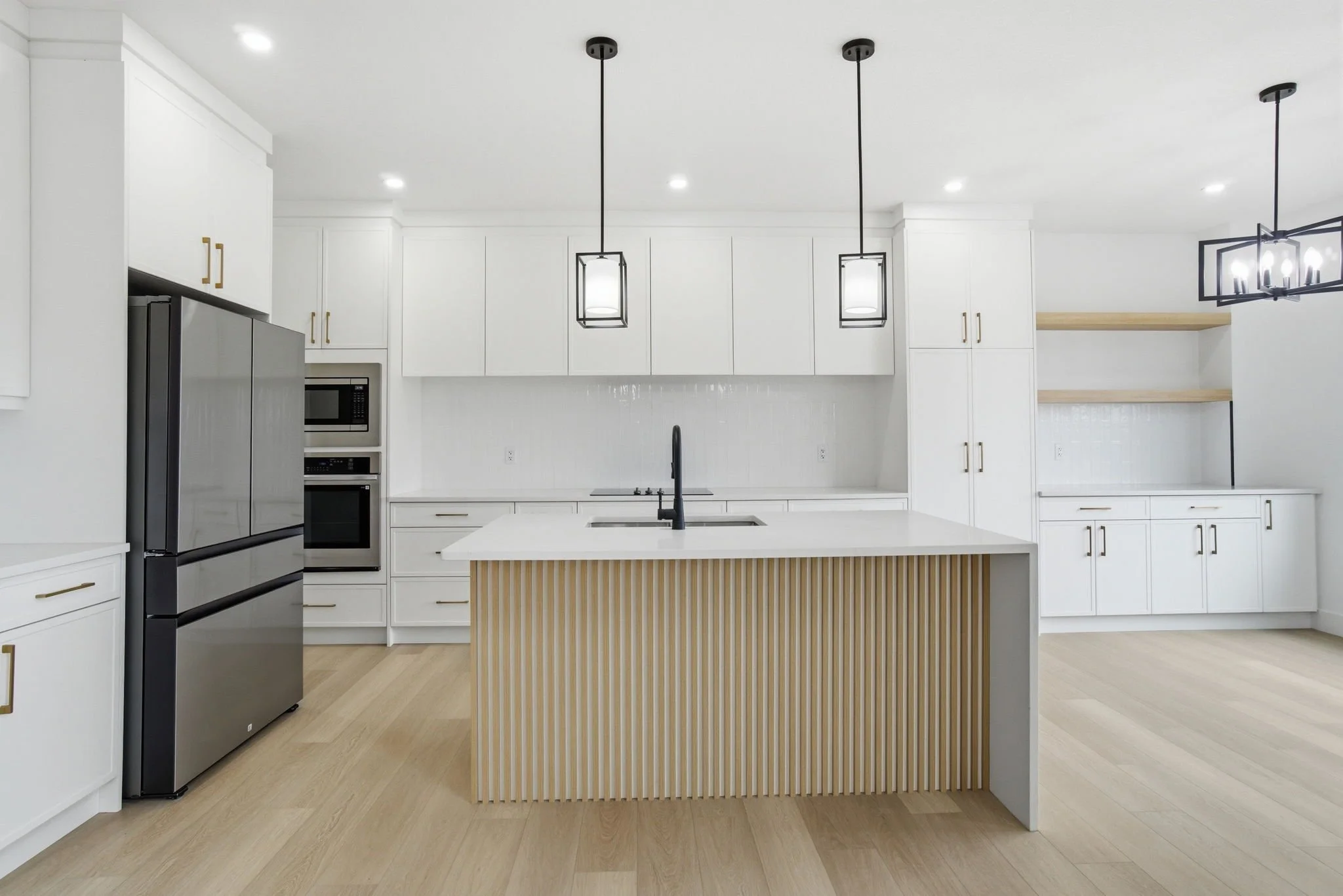 Modern kitchen with white cabinets, a central island with vertical wood slats, and black pendant and chandelier lighting fixtures.
