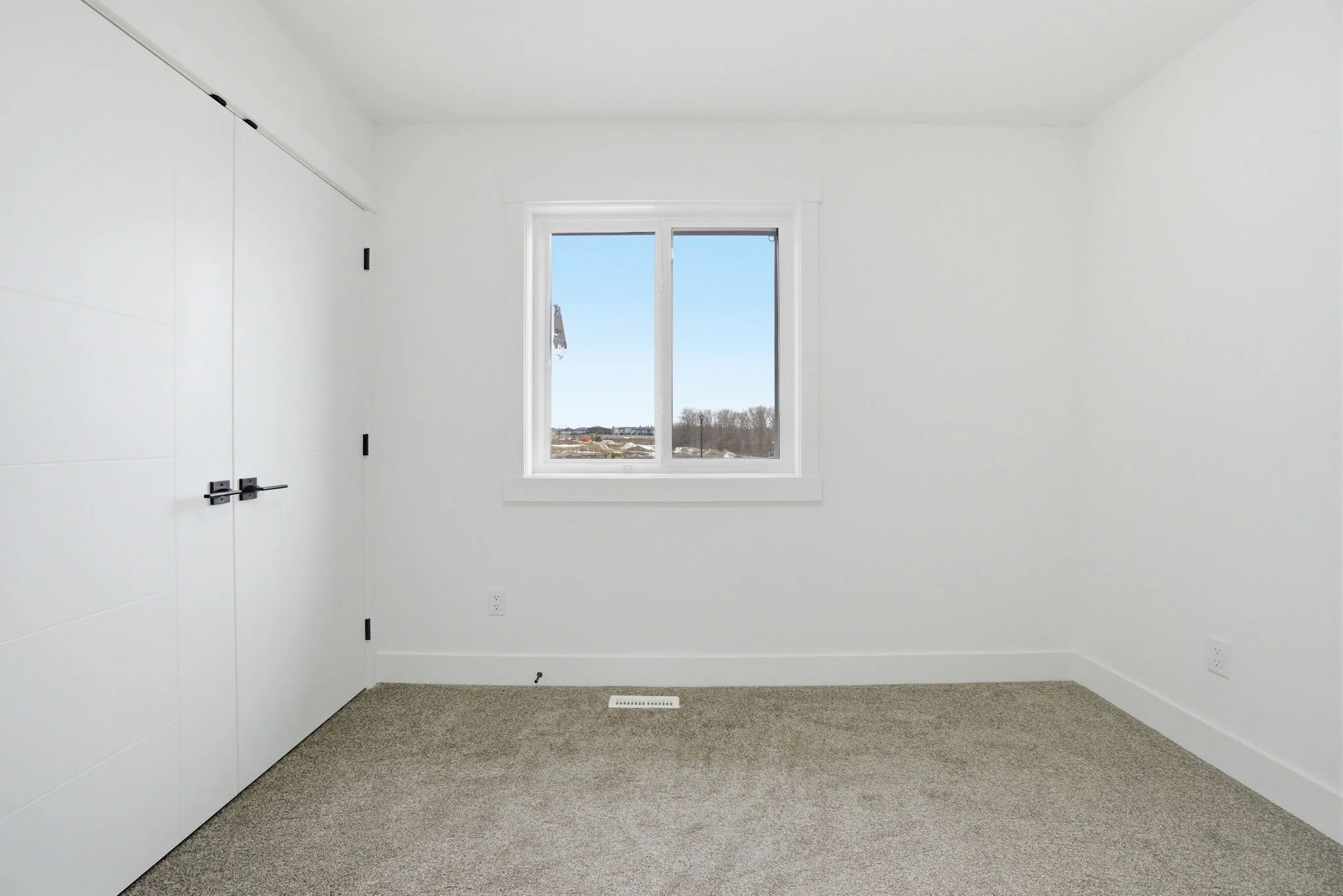 Empty room with white walls, a window showing a clear sky and construction site outside, gray carpeted floor, and closed white door with black handles.