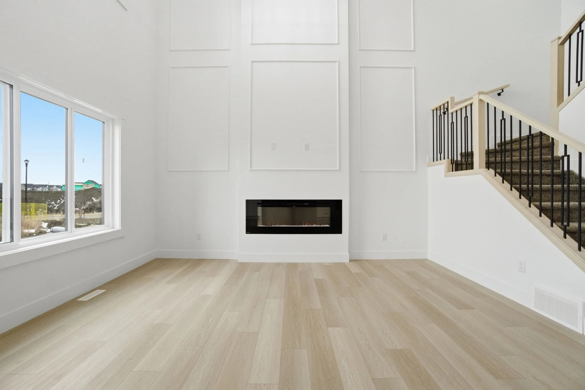 Empty living room with large window, white walls with decorative panel moldings, a modern fireplace, and a staircase with wooden handrail and black metal balusters.