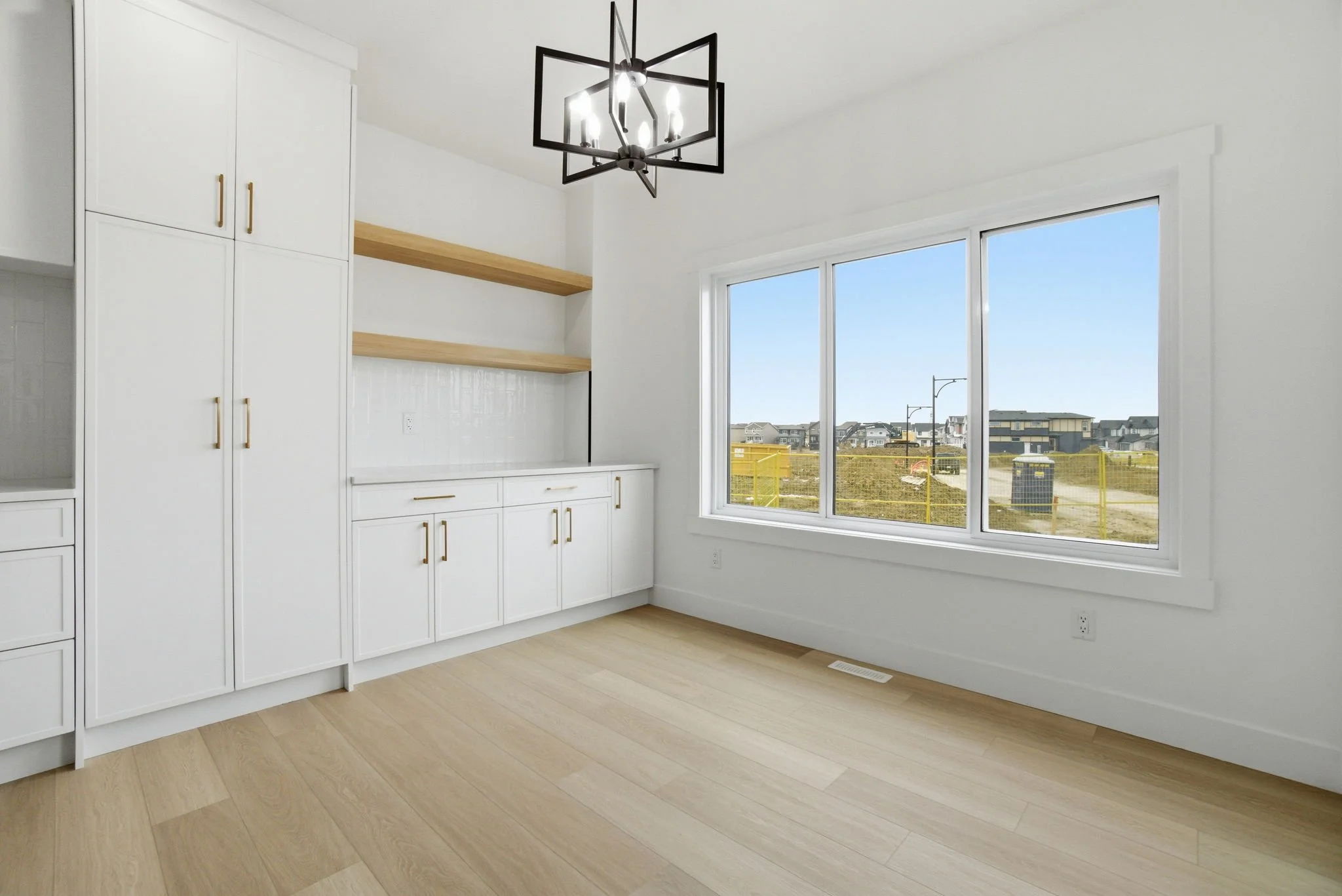 Empty room with large window showing a neighborhood outside, white walls, light wood flooring, white built-in cabinetry, and a black geometric chandelier.