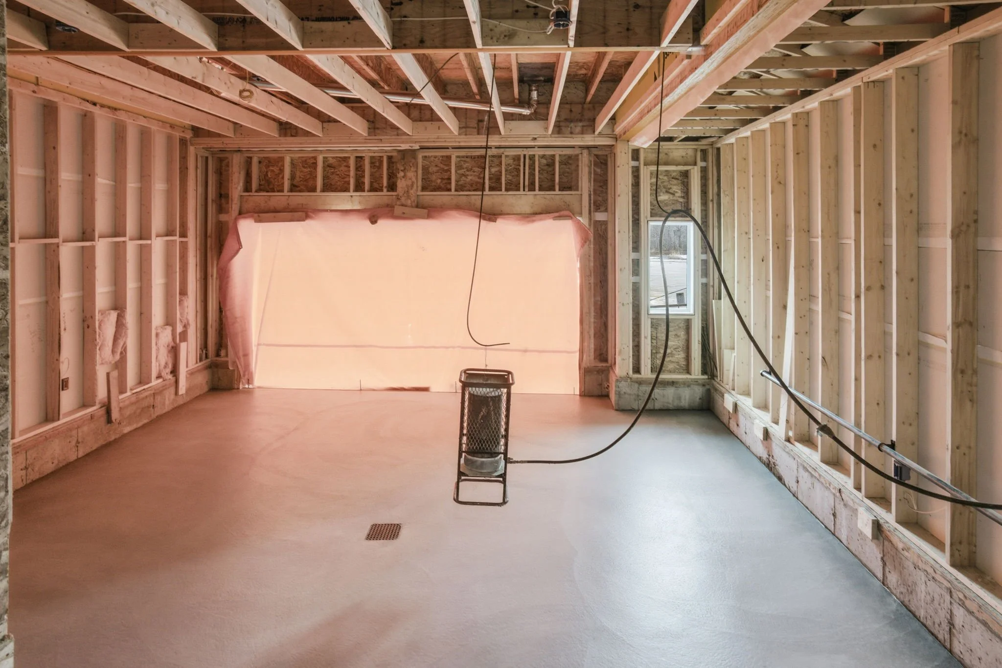 Interior view of a house under construction with exposed wooden framing, drywall visible on the ceiling, a window, and an unfinished floor with a floor drain. A small black metal chair and electrical cords are present.