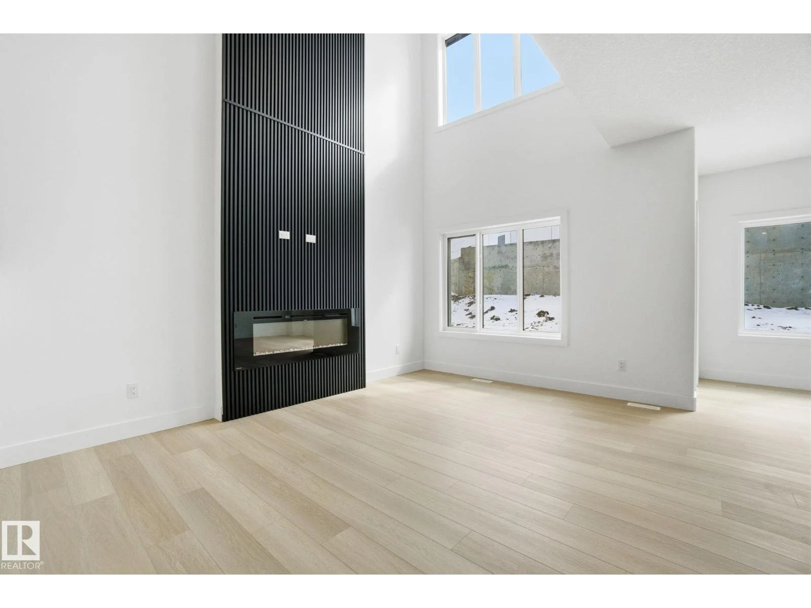 Empty living room with white walls, light wood flooring, a modern black fireplace with wood paneling, and large windows showing outdoor snow.