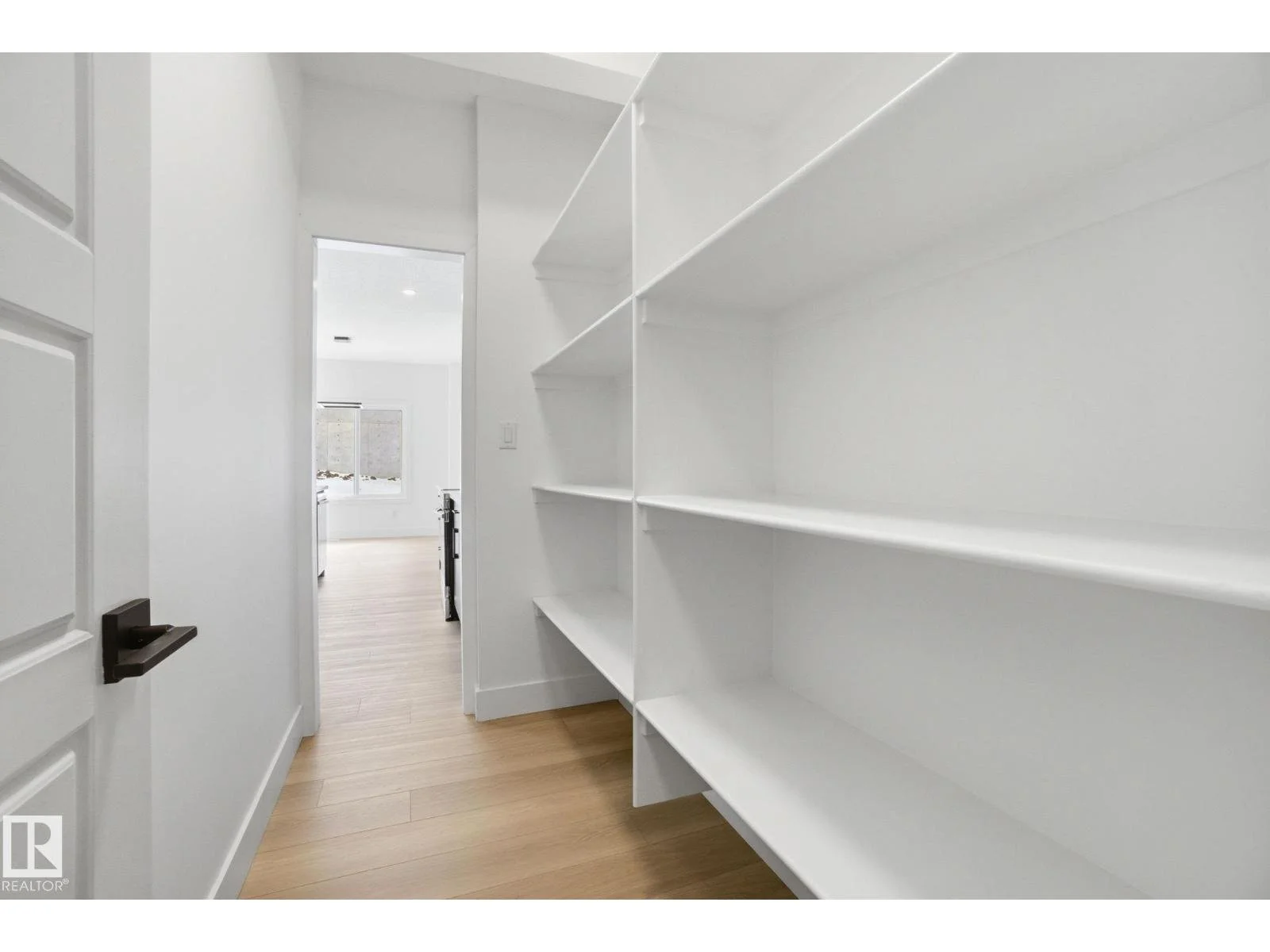 White built-in shelves in a narrow pantry or storage room with a view into a bright kitchen area