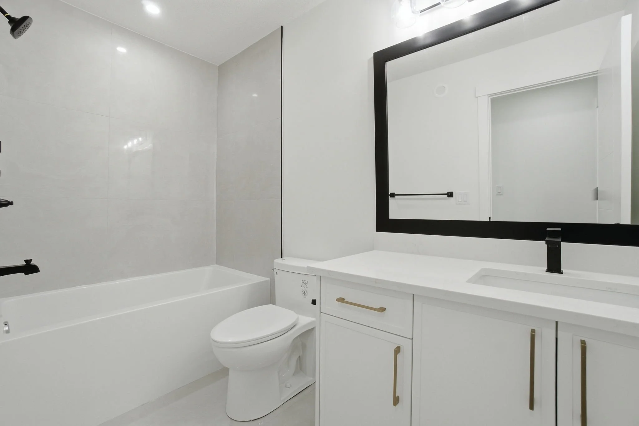 A modern, minimalist bathroom with white walls, black fixtures, and a large mirror above a white vanity. Includes a bathtub, toilet, and a black towel bar.
