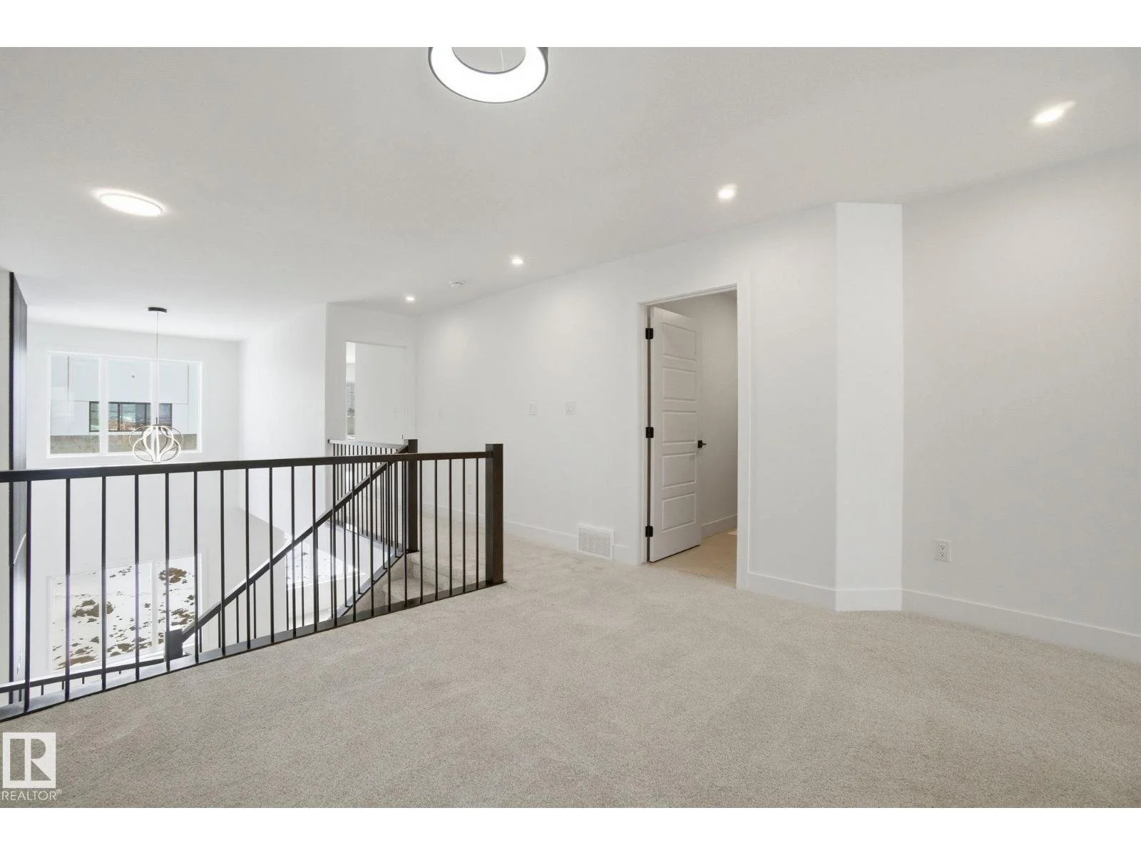 Bright, modern, white-painted interior of a home upstairs landing area, with a staircase leading down, and an open space with ceiling lights and a window.