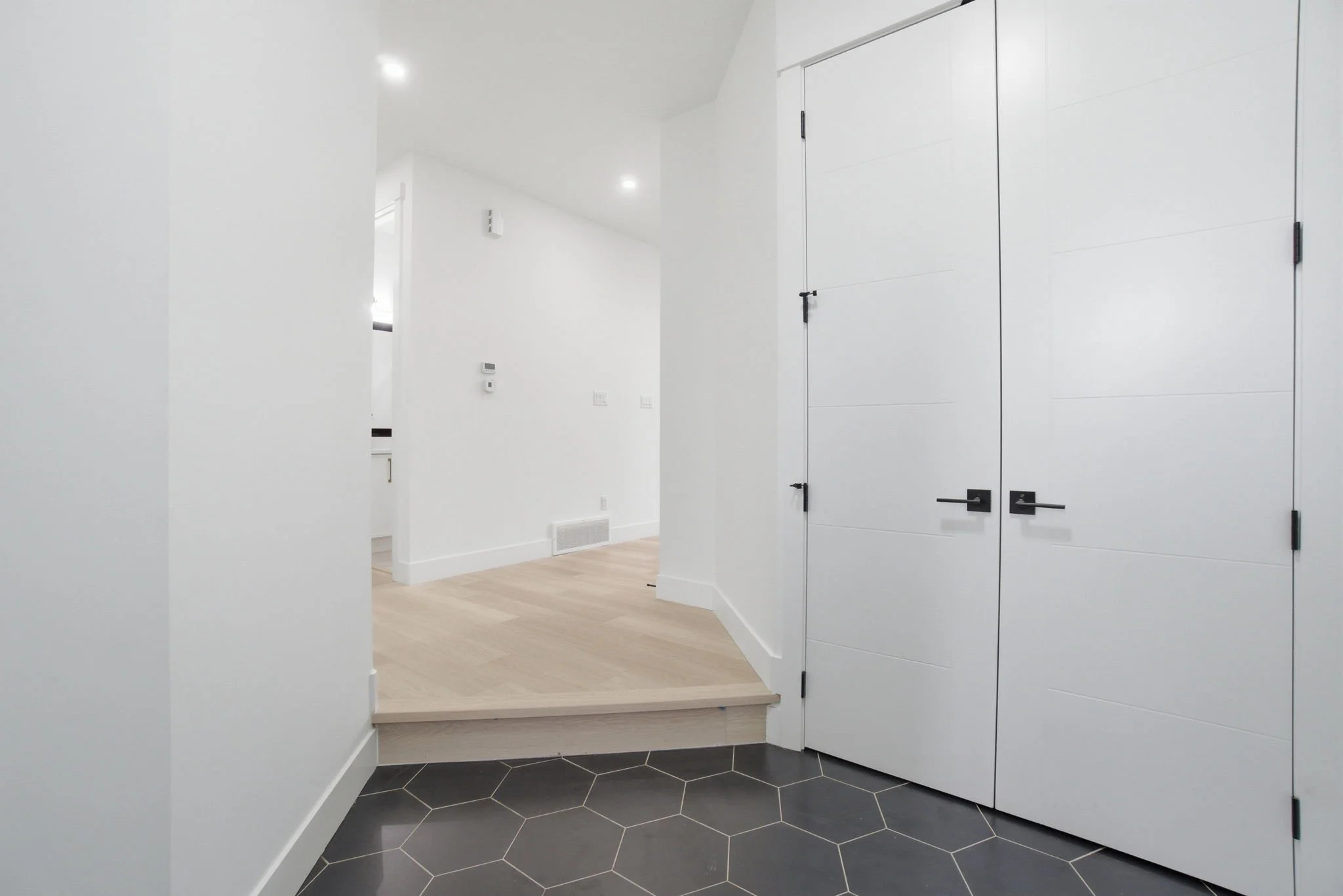 Interior view of a modern home entryway with black hexagonal floor tiles, white walls, and built-in white storage cabinets with black handles.