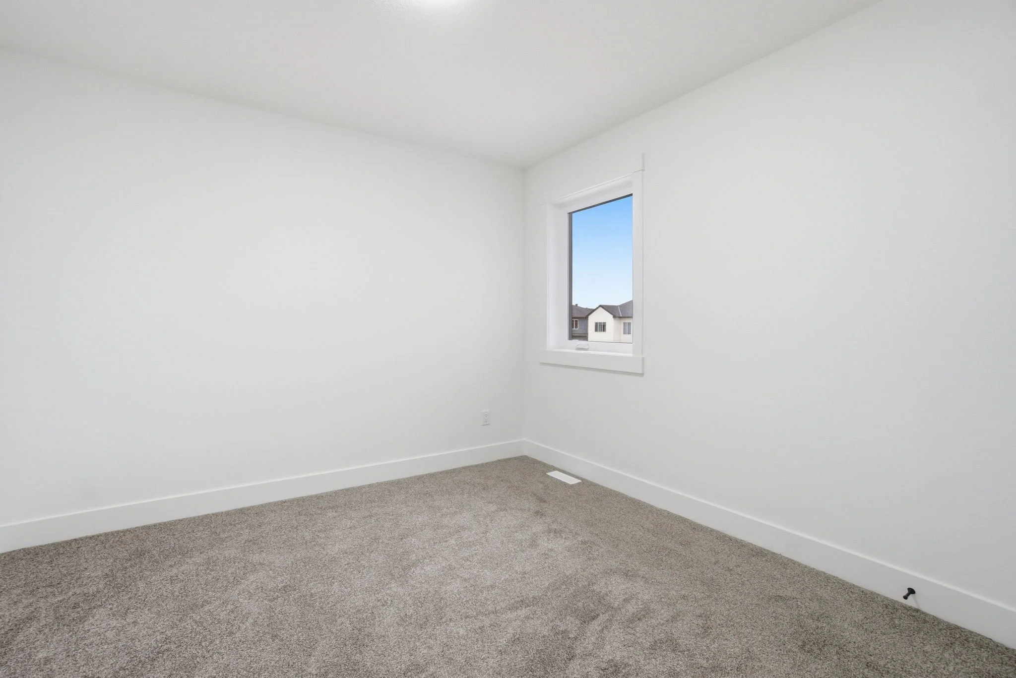 Empty white room with a window showing houses outside and beige carpeted floor.