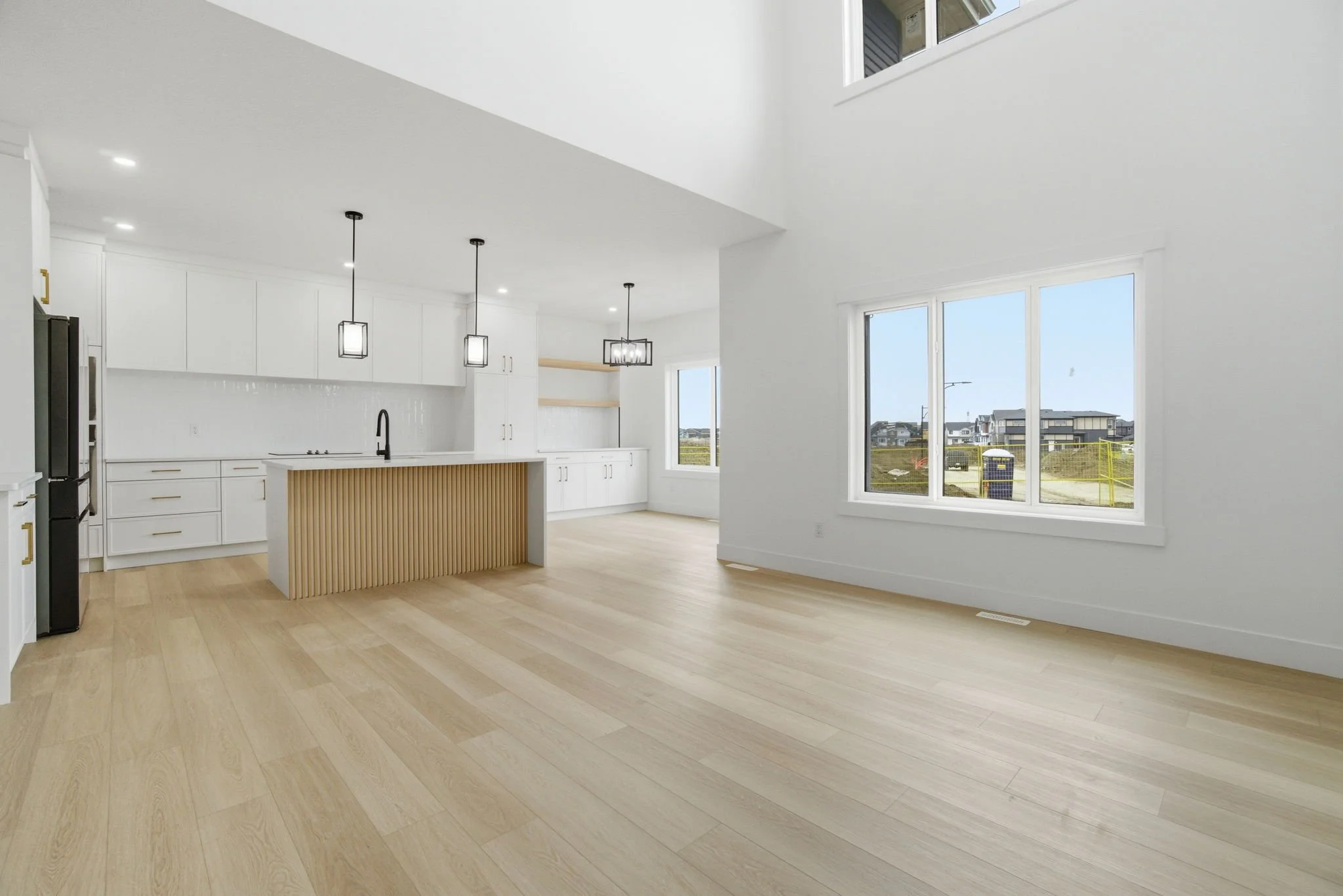 Interior view of a modern, spacious, white kitchen and living area with large windows, light wood flooring, and minimalist pendant lighting.