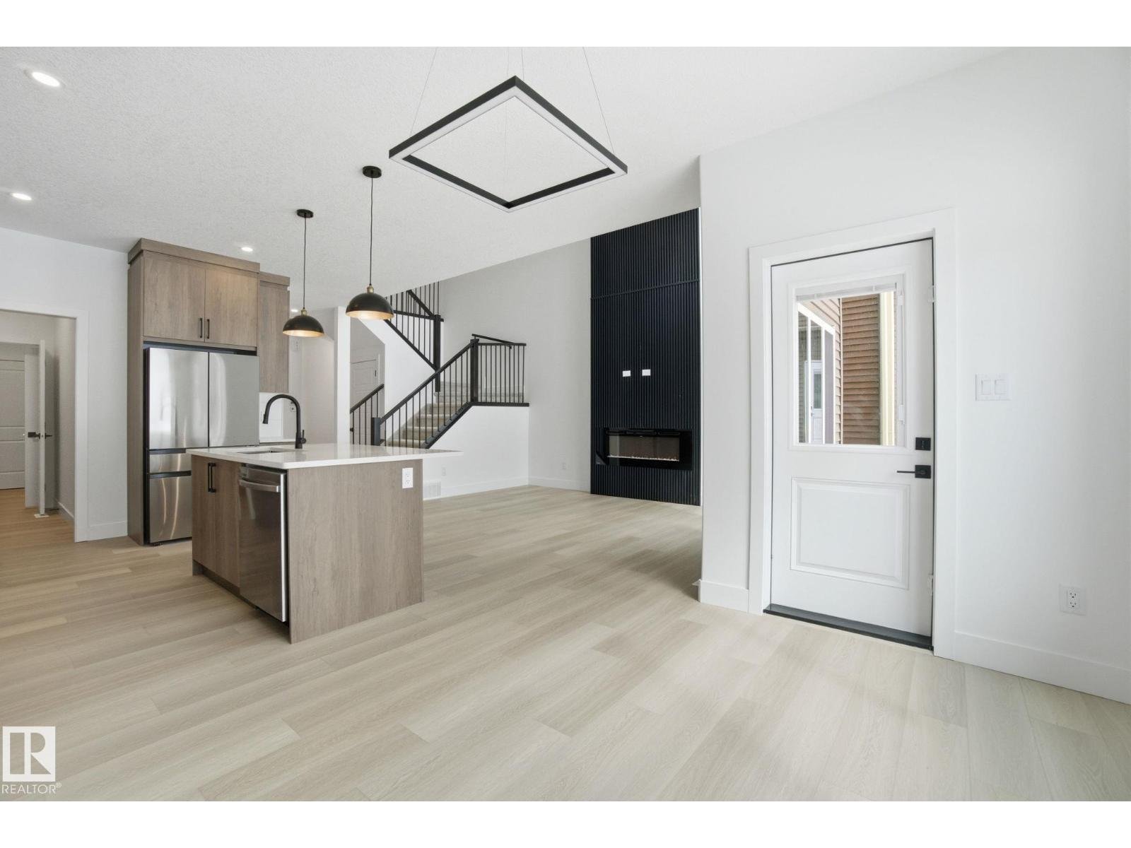 Modern open-concept living space with kitchen featuring light wood cabinets, stainless steel appliances, island with sink, black pendant lights, stairs with black railing leading upstairs, black textured wall with fireplace, and a white door with win