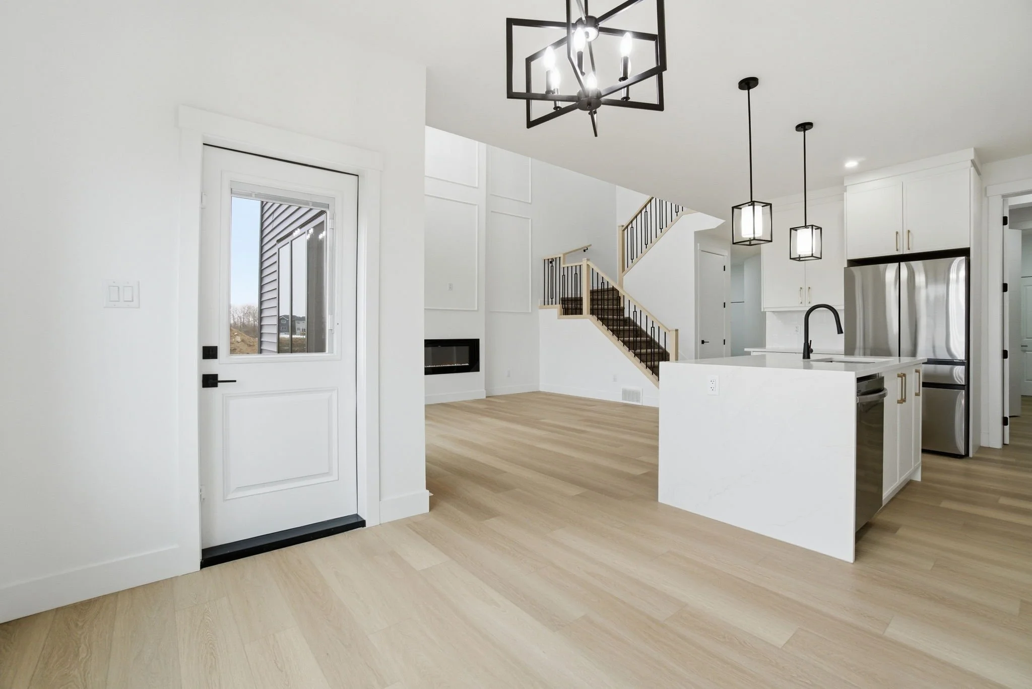 Modern kitchen and living room with white walls, light wood flooring, black and gold fixtures, stainless steel appliances, large windows, staircase, and minimal decor.