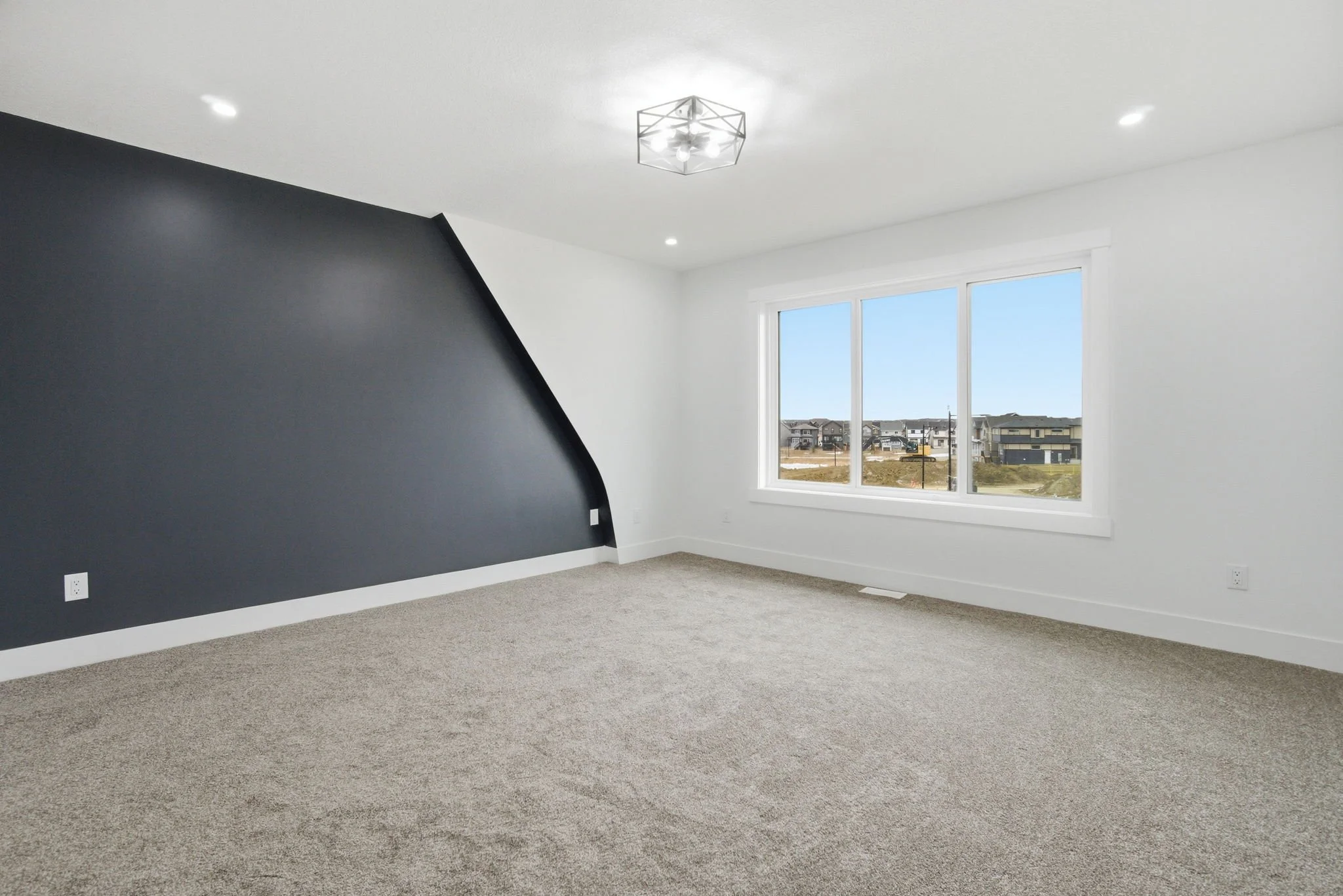 Empty room with gray carpet, white walls, a dark gray accent wall, large window showing neighborhood houses, ceiling light fixture, and recessed lighting.