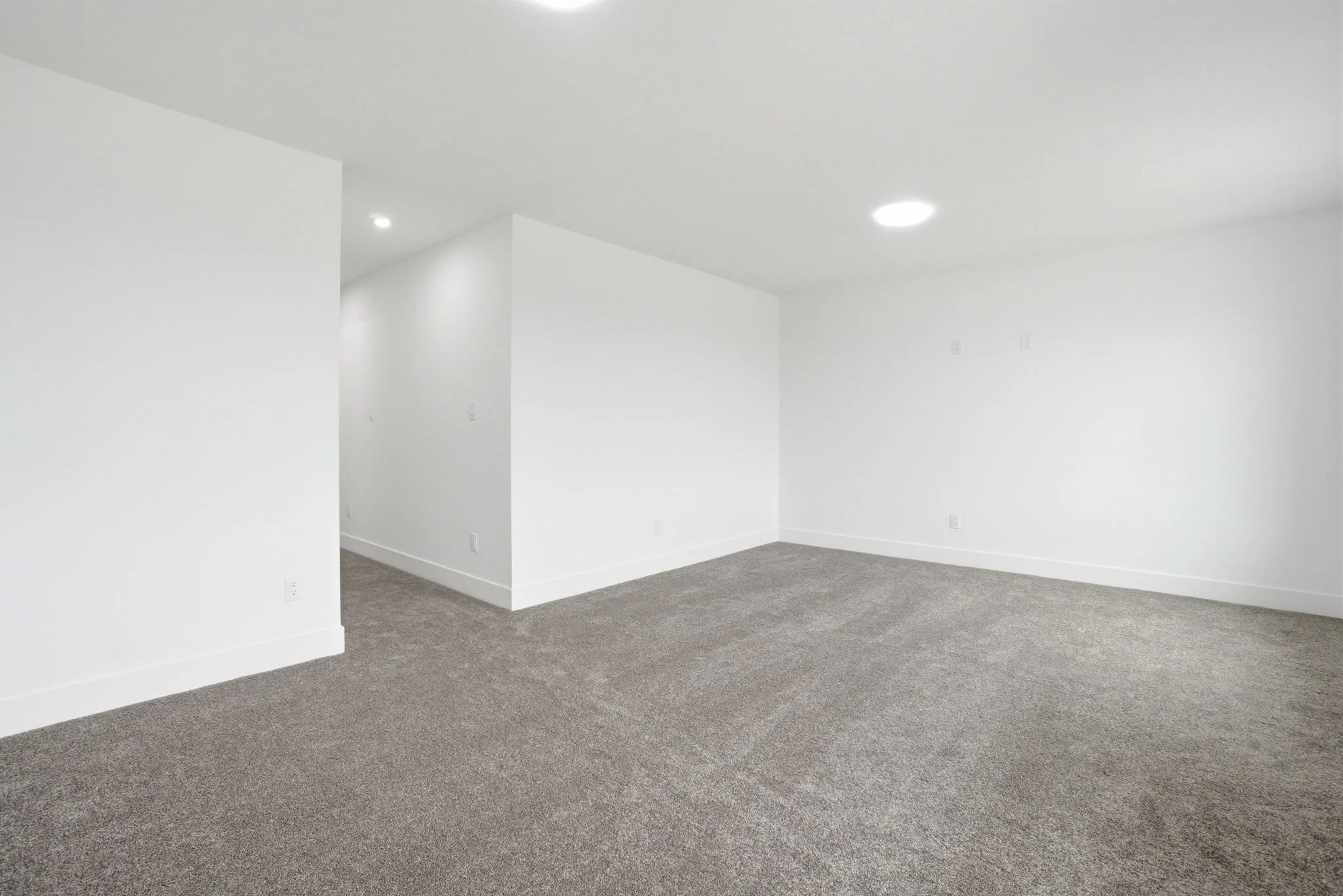 Empty white room with gray carpet flooring and recessed ceiling lights.
