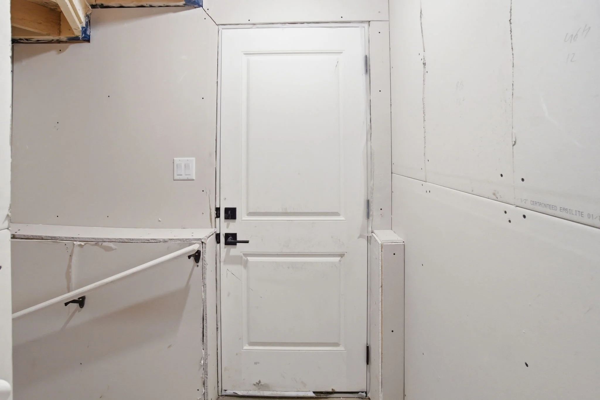 Interior view of a room under construction with white drywall walls, a white door with molding, electrical outlets, and exposed framing in the upper left corner.