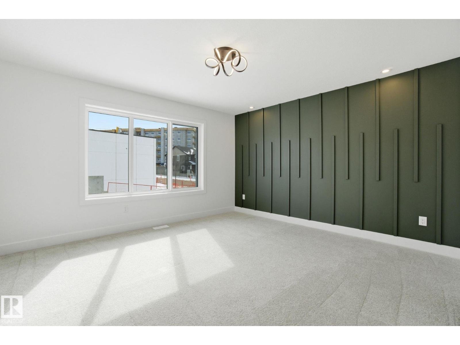 Empty modern room with a large window, white walls, a green accent wall with vertical paneling, beige carpet, ceiling light fixture, and daylight streaming in from outside.