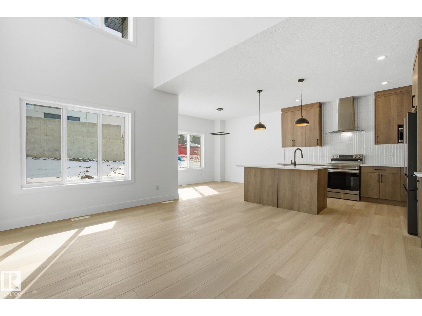 Interior view of a modern, empty kitchen with light wood flooring, white walls, large windows, and natural light. The kitchen has wooden cabinets, a kitchen island with a black faucet, and stainless steel appliances. There are ceiling lights and pend