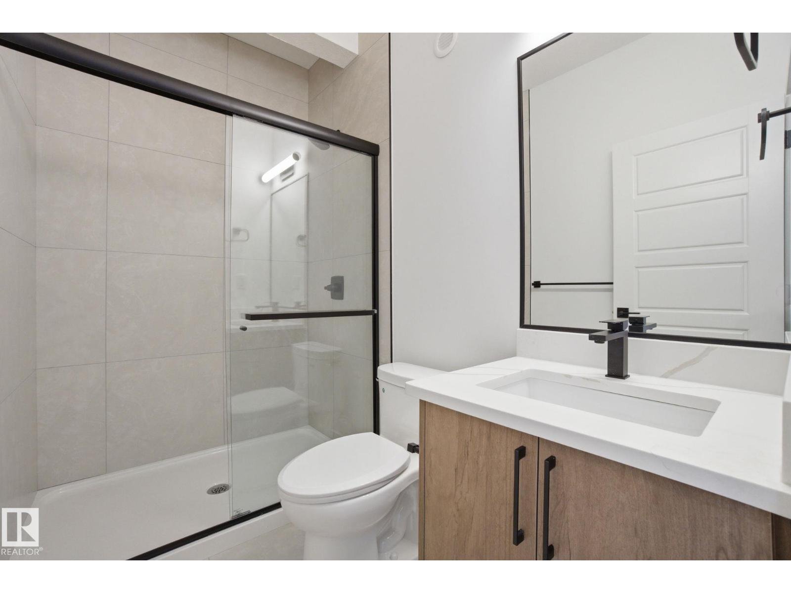 Modern bathroom with glass-enclosed shower, white toilet, wooden vanity with black hardware, white countertop, large mirror, black faucet, and a white door.