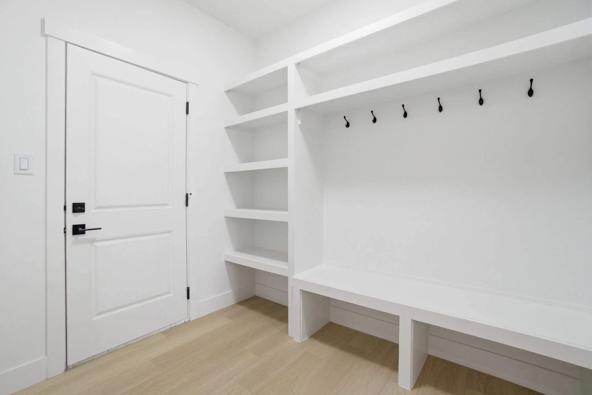 Empty white entryway with built-in shelves and hooks on the wall, near a closed white door with black hardware and a light switch.