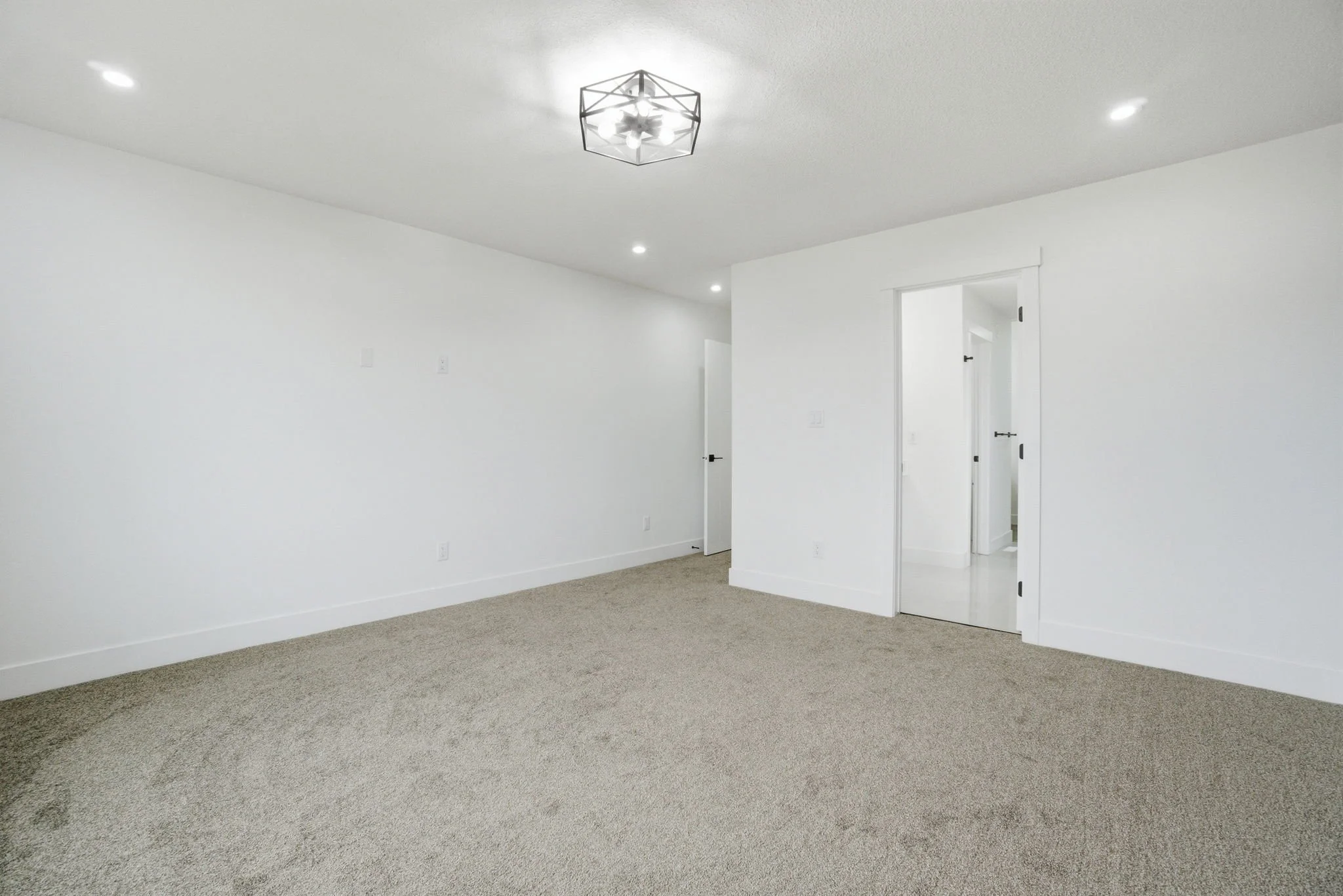 Empty white room with beige carpet, open door, and ceiling light fixtures.
