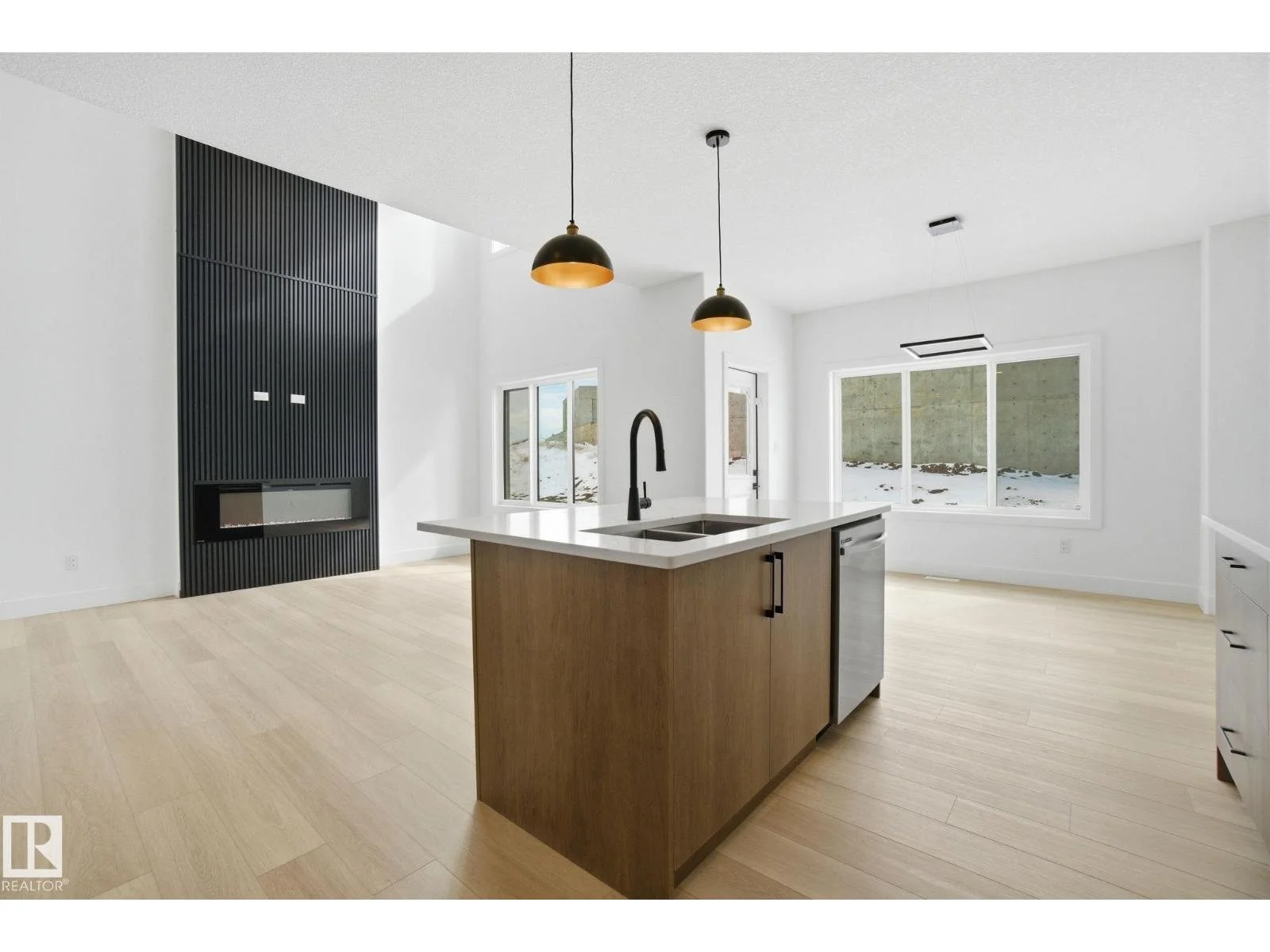 Modern kitchen with white walls, light wood flooring, a kitchen island with a white countertop, black faucet, and stainless steel dishwasher, large windows, and pendant lights.
