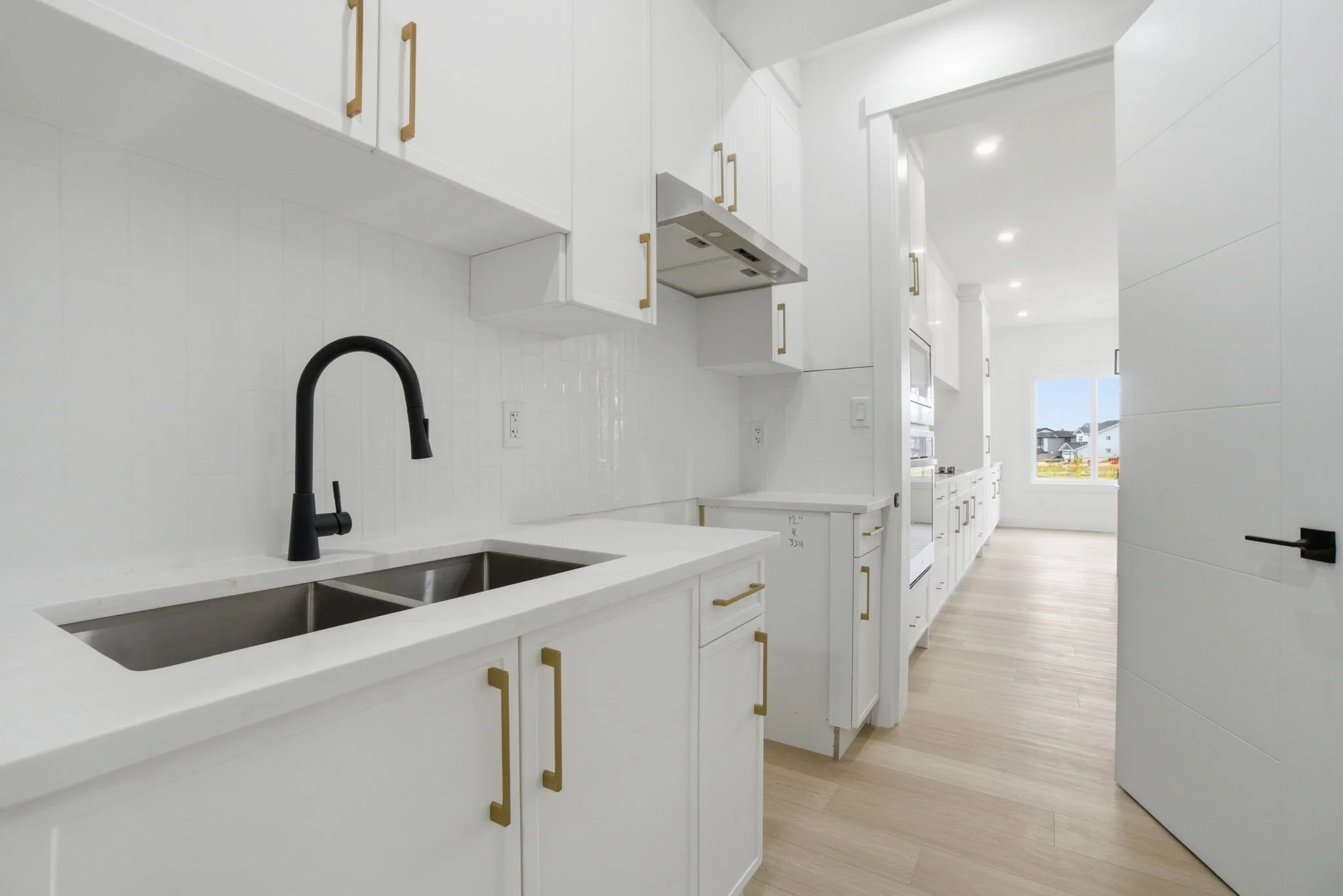 Modern white kitchen with gold handles, black faucet, and large window showing a neighborhood outside.