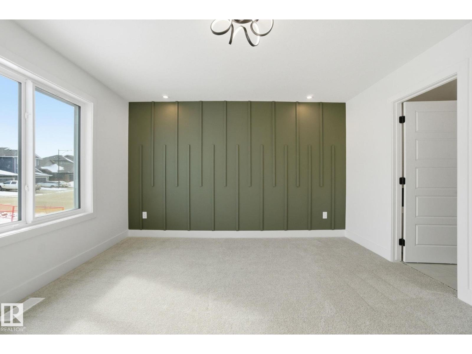 Empty room with large window, white walls, beige carpet, and a green accent wall with vertical panels.