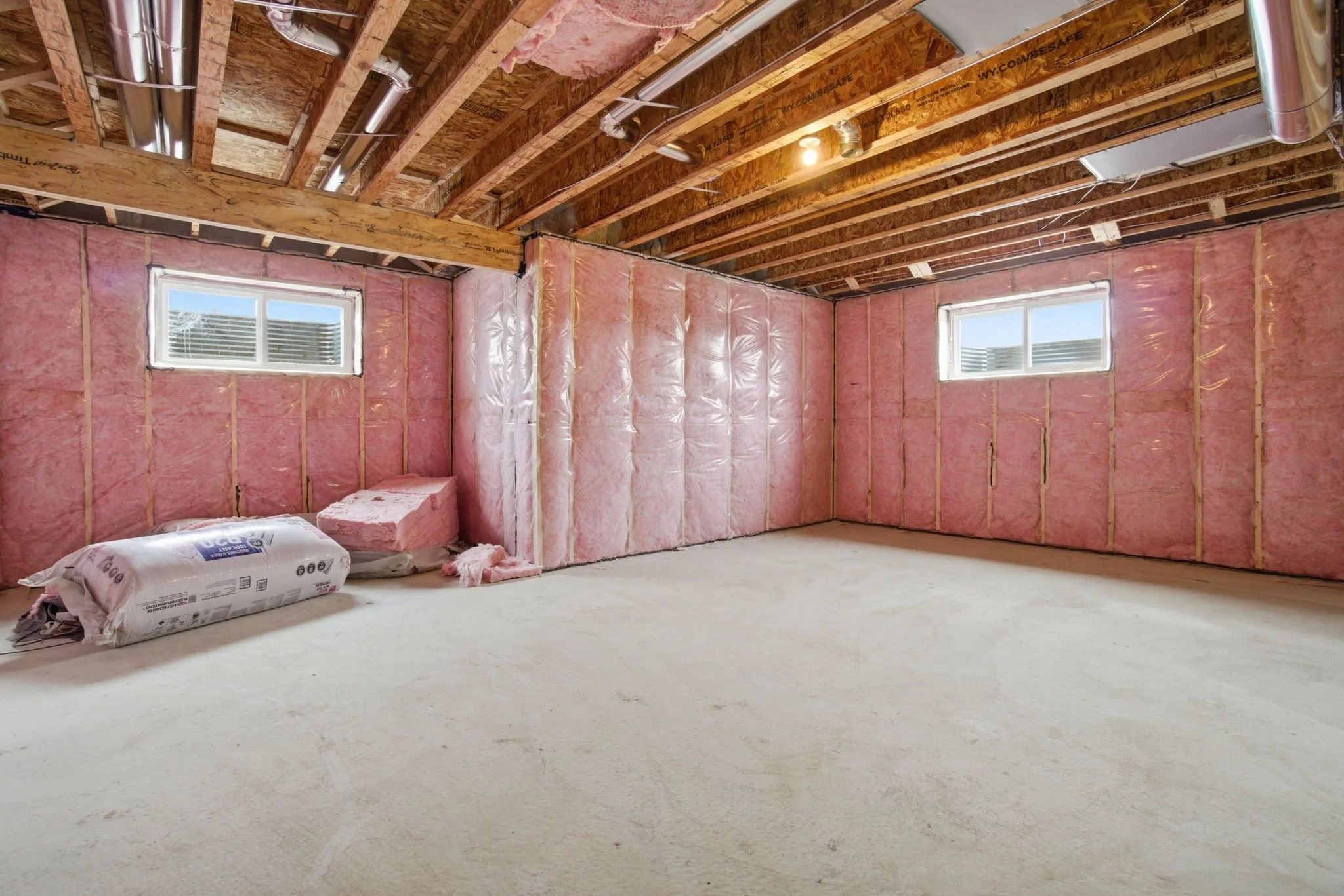 A partially finished room under construction with pink insulation on the walls, exposed ceiling beams, and ducts running near the ceiling. There are two small windows letting in natural light and a bag of insulation material on the floor.