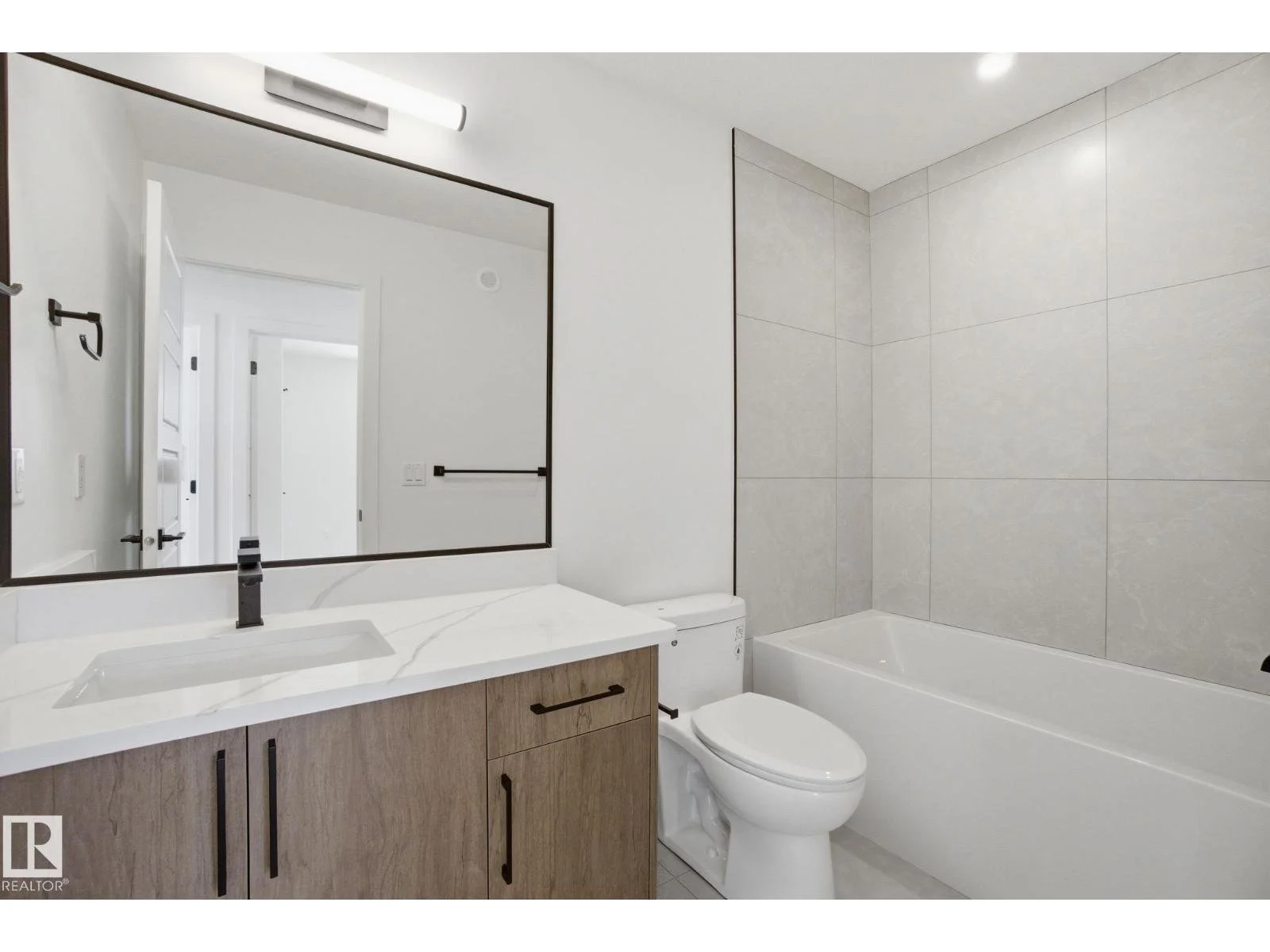 Modern bathroom with a large mirror, white marble countertop, black fixtures, light wood vanity, toilet, bathtub, and light gray tiled walls.