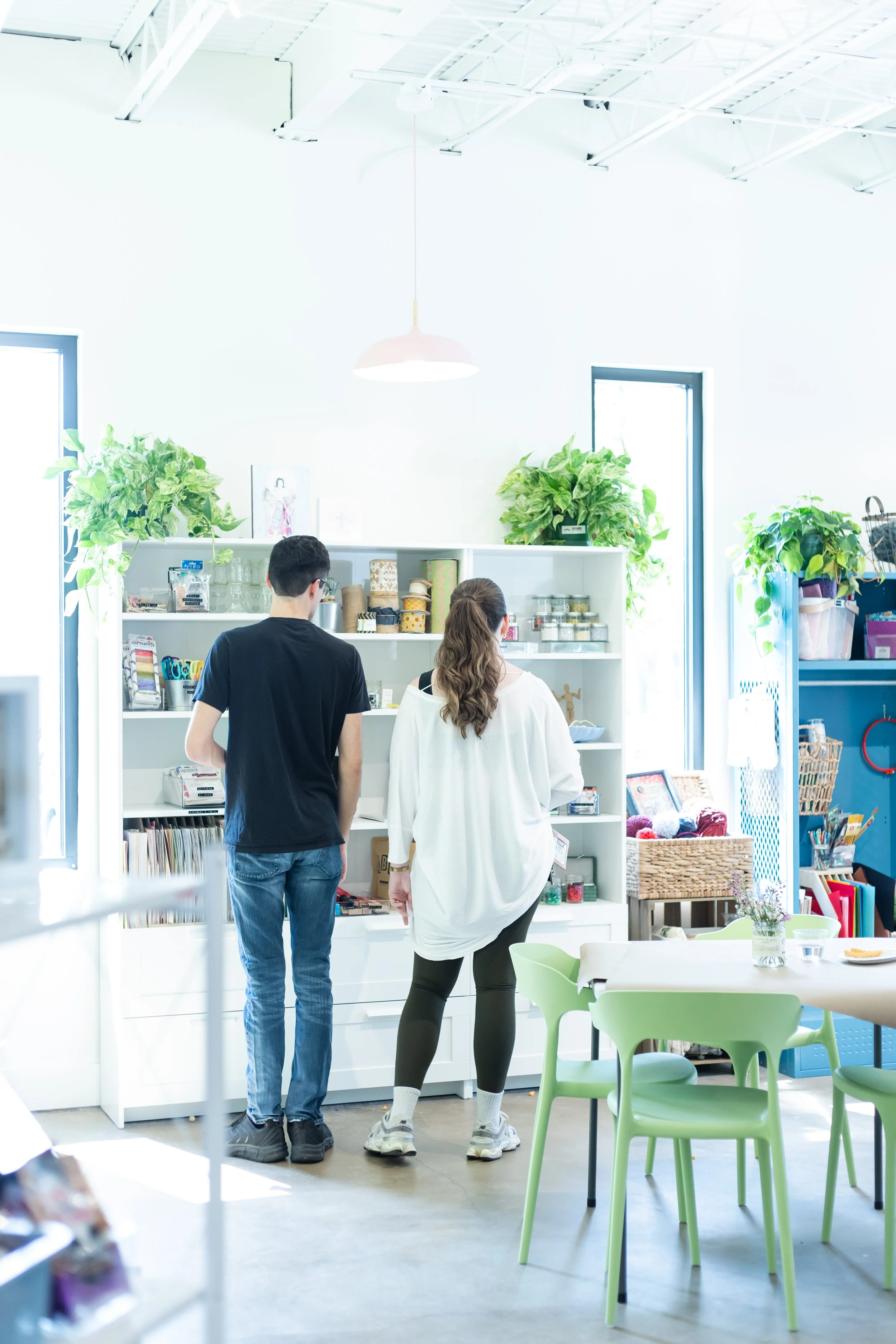 A man and woman studying The Craft Room with shelves containing scrapbooking paper, stickers, jewelry making supplies, beads, and found objects. A light and bright space with greenery and lots of natural light.