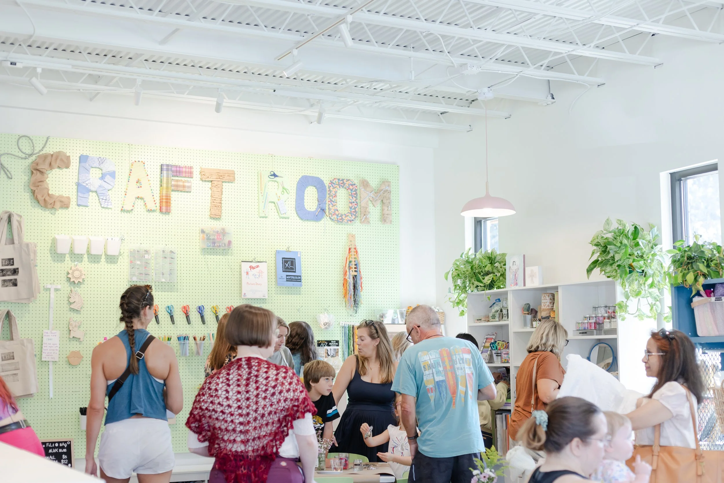 Wide shot of The Craft Room including a crowd of people shopping and  a handmade sign in the background made out of recycled fabric, arts and craft supplies, and leftover shipping and construction materials. 