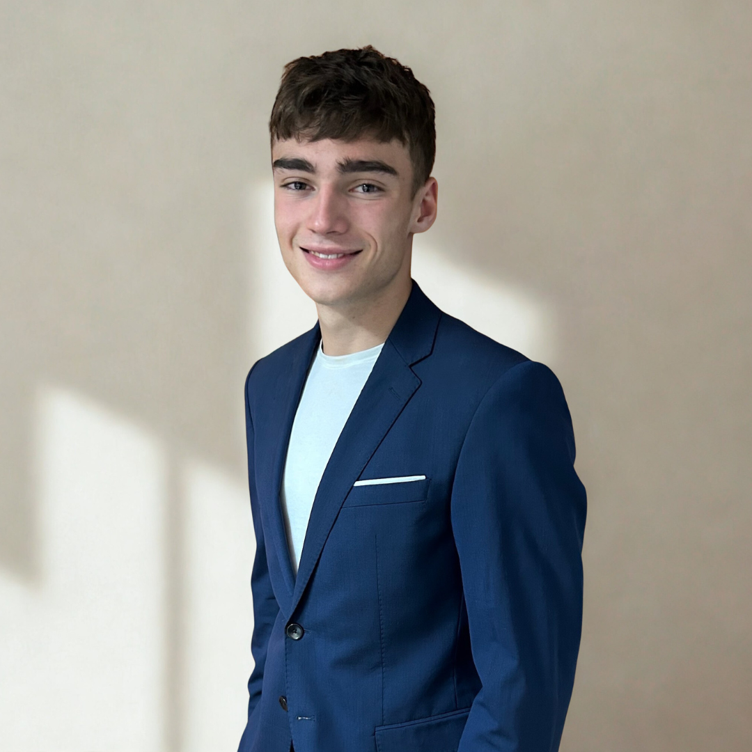 A young man with short brown hair, wearing a navy blazer and a white shirt, standing against a plain light-colored background.