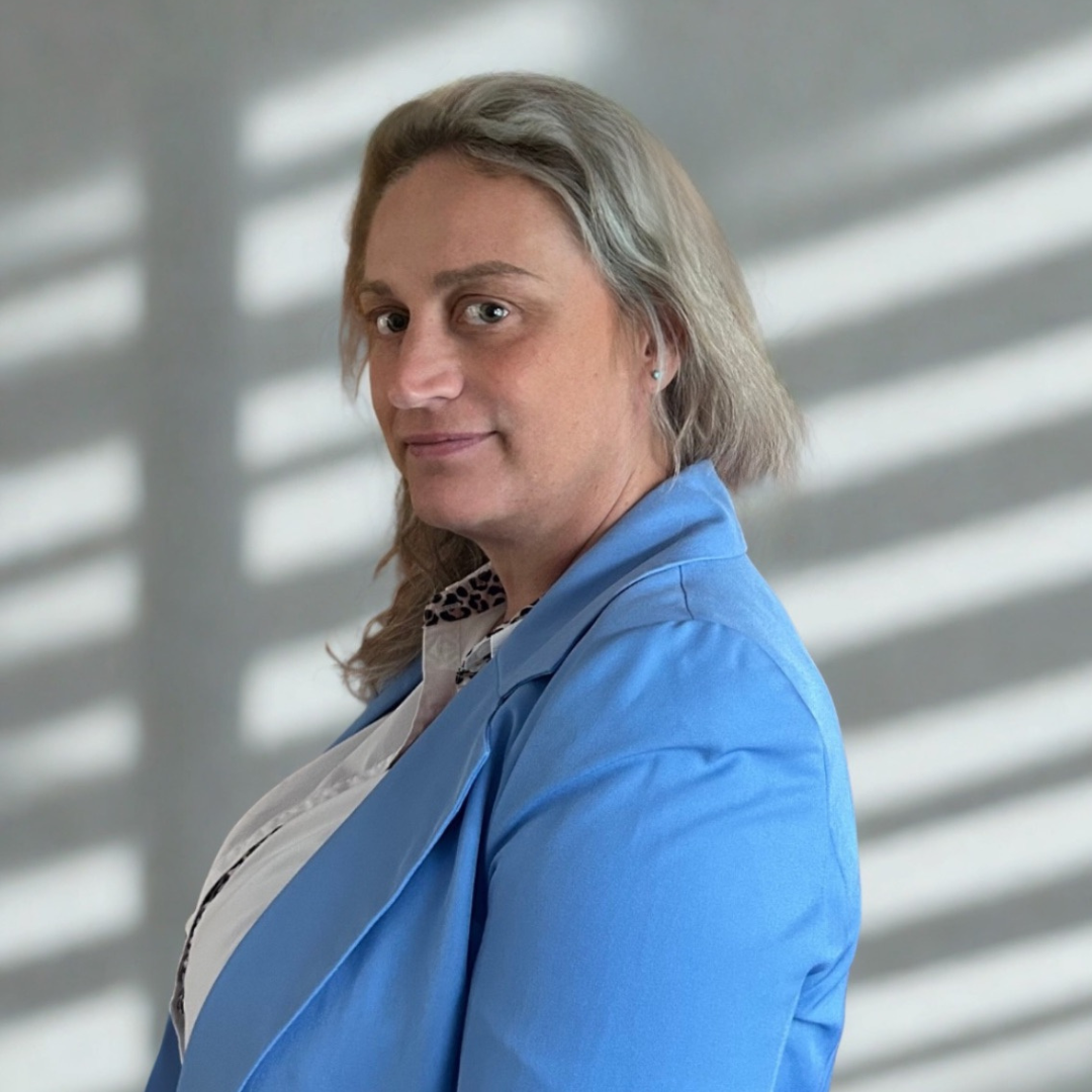 A person with shoulder-length gray hair, wearing a blue blazer and white shirt, standing against a light background with shadows.