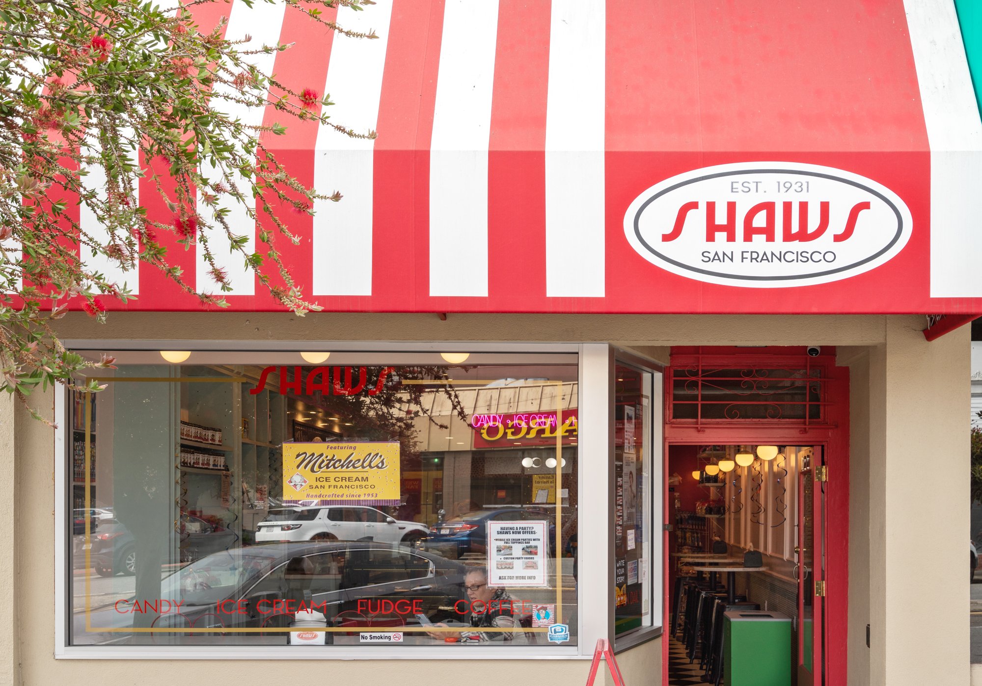 Shaw's San Francisco storefront with red and white striped awning, featuring signs for candy, ice cream, and fudge. The window displays a Mitchell's Ice Cream poster and inside view of the store.