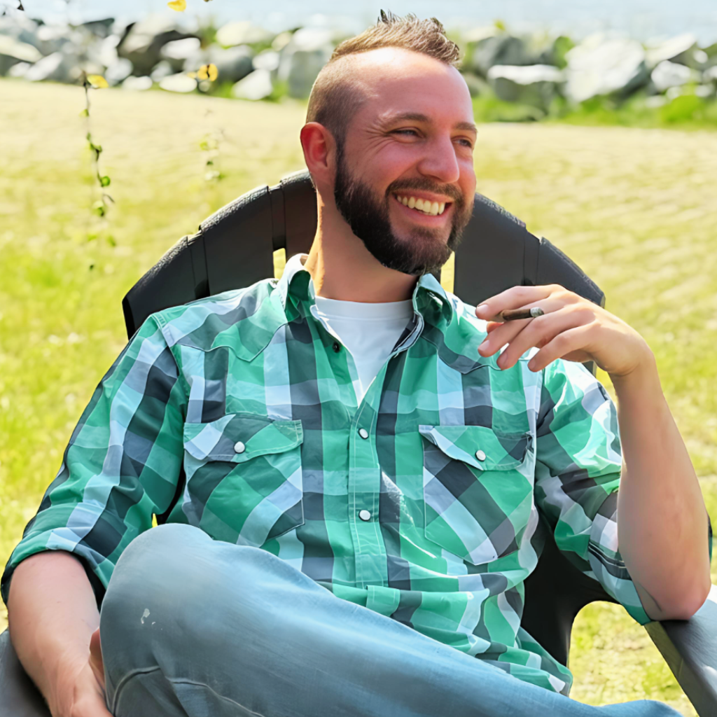 A smiling man with a beard and a plaid green shirt sitting outdoors on a lawn chair in a field, holding a cigar in his hand.