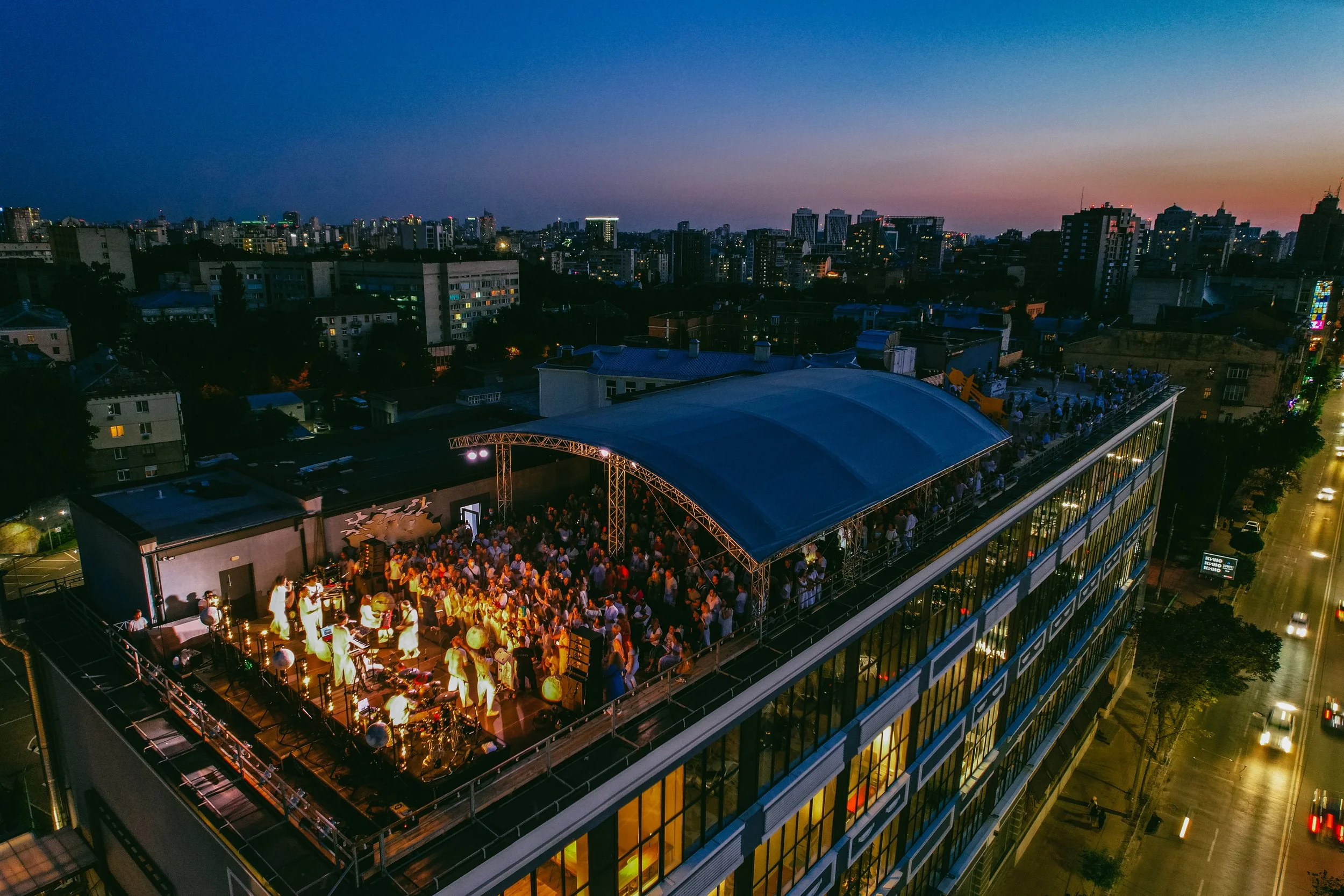 An aerial view of a large rooftop party on a building in a city at dusk, with a crowd dancing and a band performing under a curved roof structure, and the city skyline in the background.