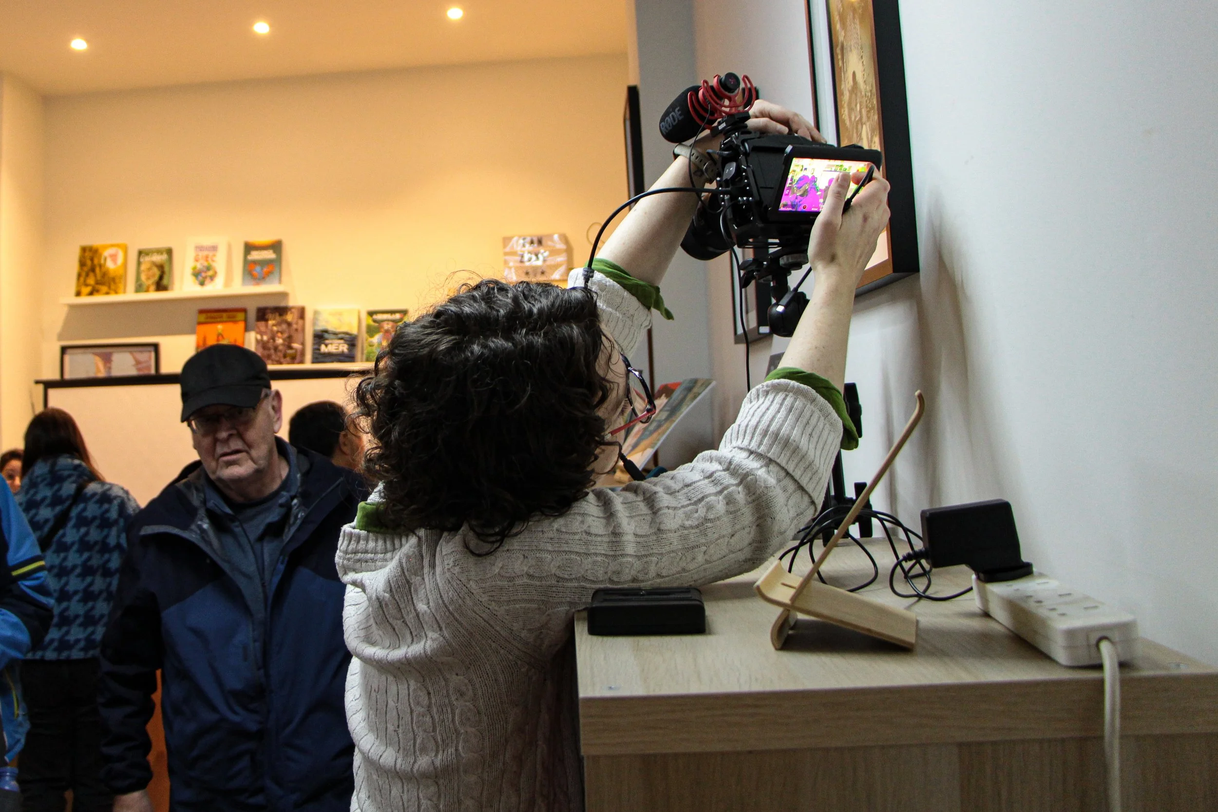 Aoife Buggy is setting up a camera in a production environment to record a podcast in Edinburgh Scotland, operating the camera, microphone and lights. Audience in the background, in a room with framed pictures and books on a shelf for podcast.