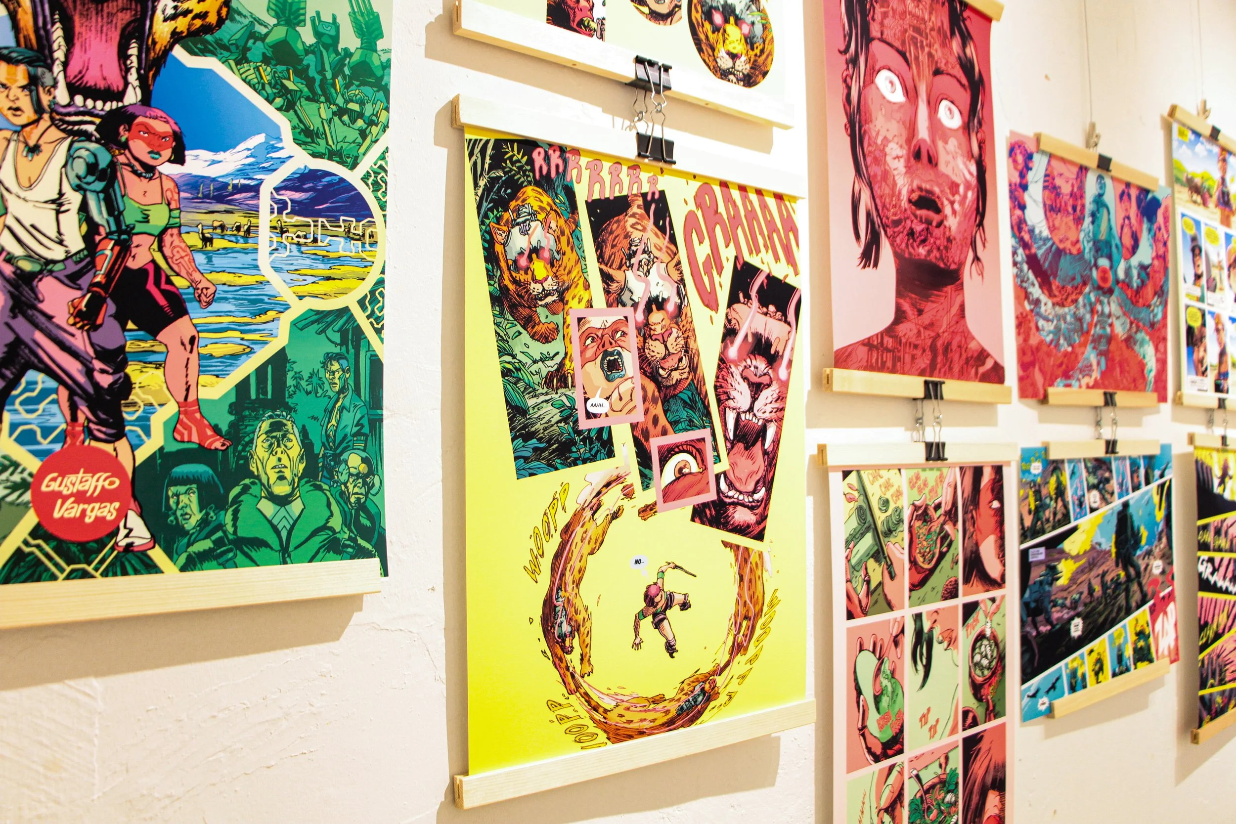 Part of a comic book art display with colorful illustrations of characters and scenes, including a woman with a gun, a tiger, and a person with a snake, featuring bold and vibrant colors.