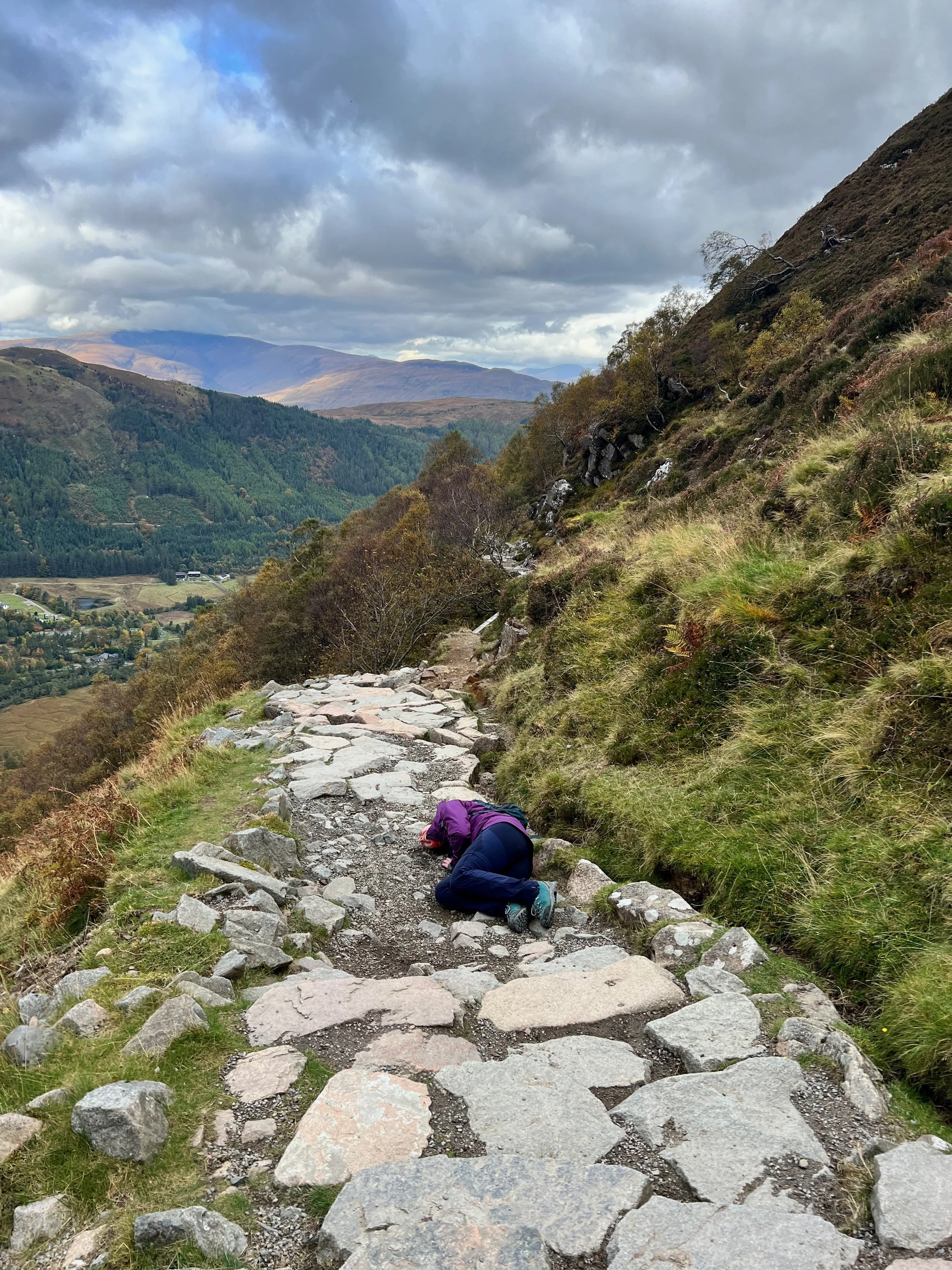 Aoife Buggy, a freelance video editor and motion graphic designer, stumbles on a path while hiking on a rocky trail in a mountainous landscape with green hills near Edinburgh, Scotland