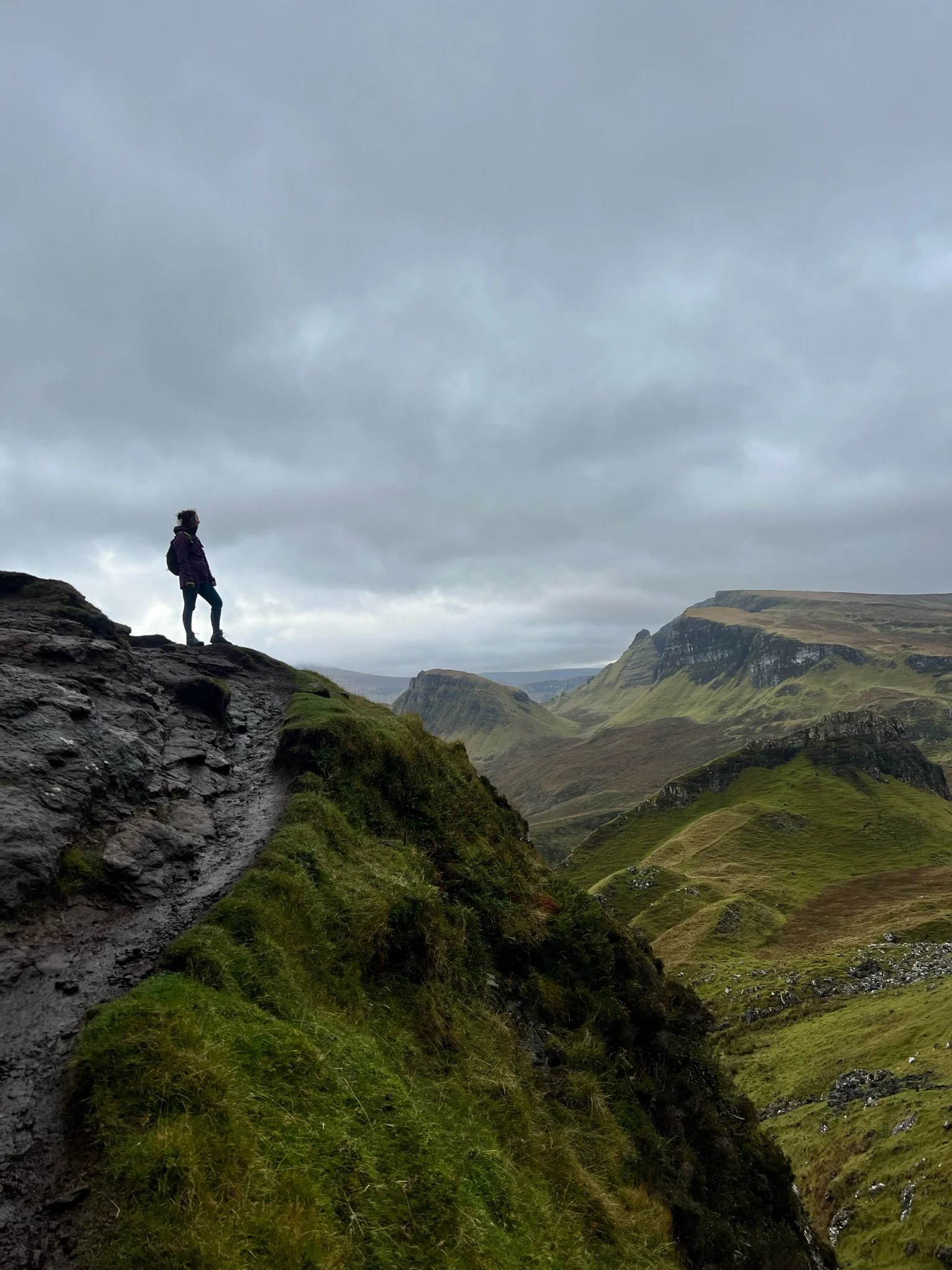 Person standing on a rocky cliff edge overlooking a vast green valley with rolling hills under a cloudy sky.