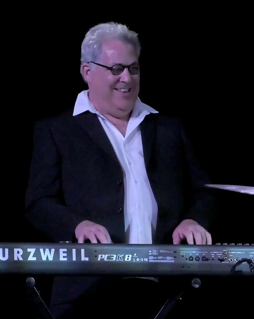 A man with gray hair, glasses, and a broad smile playing a Kurzweil keyboard on a dark stage.
