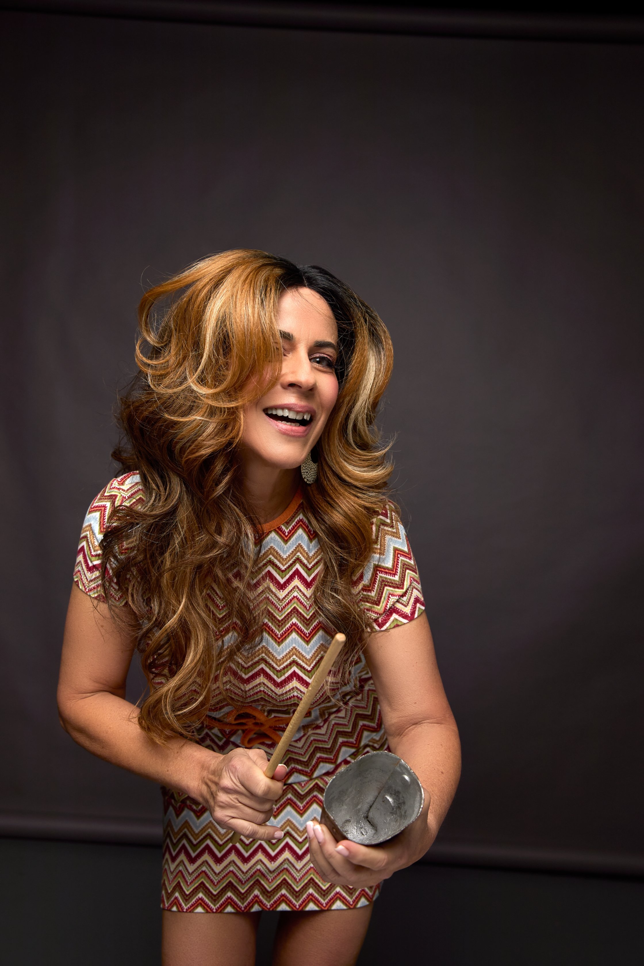 A woman with long, wavy, light brown hair smiling while holding a percussion instrument and a drumstick, wearing a colorful, zigzag patterned dress.