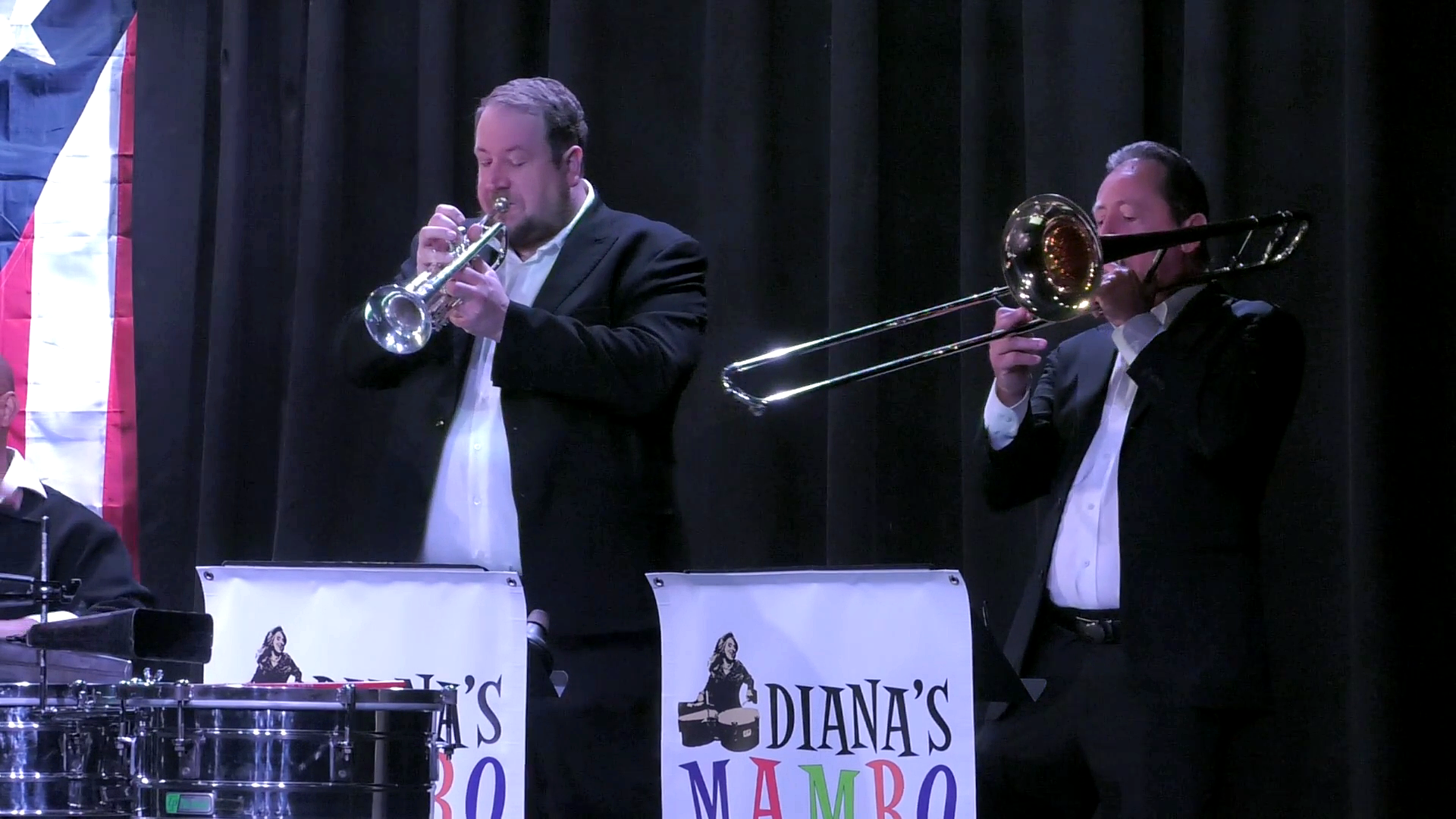 Two musicians in black suits playing brass instruments, one with a trumpet and the other with a trombone, on a stage with black curtains and signs that read "Diana's Mambo."