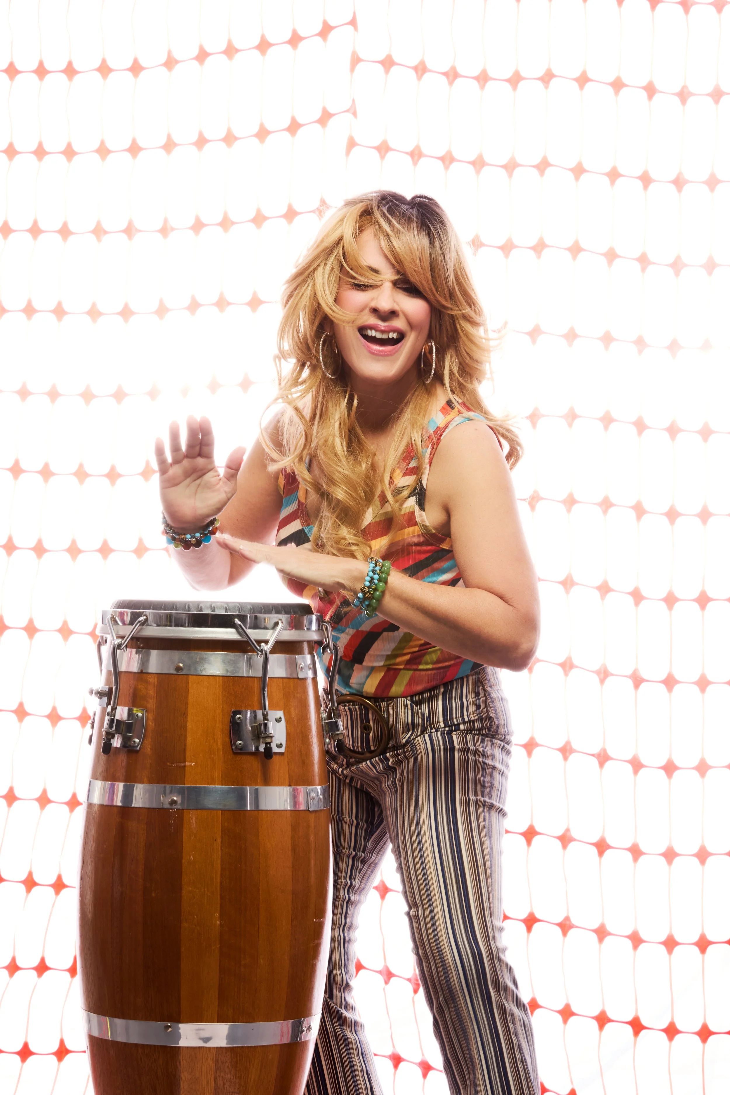 A woman with long blonde hair wearing a colorful sleeveless top and striped pants, playing a conga drum with her hands raised, smiling and looking joyful against a background of orange construction fencing.