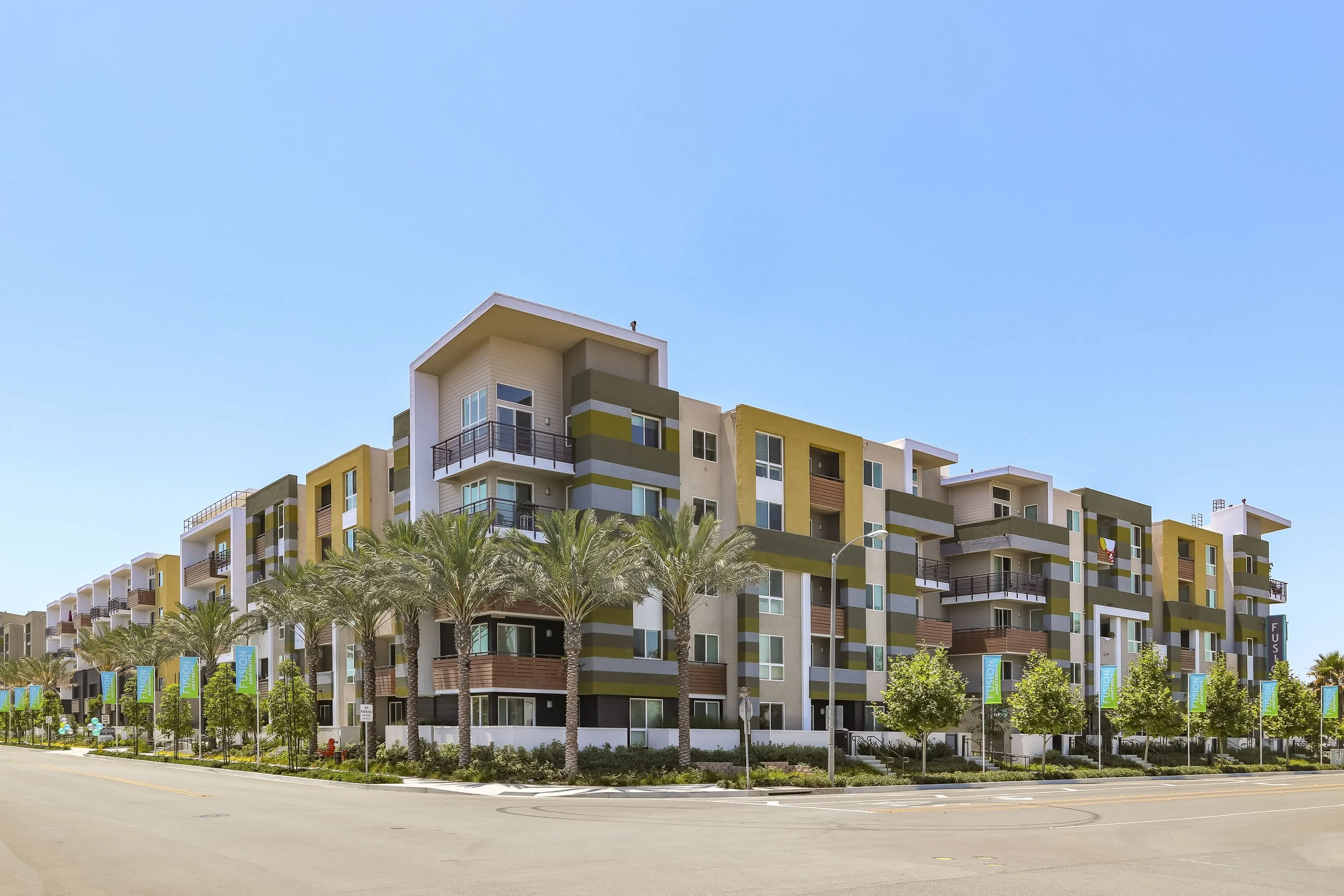 Multifamily apartment buildings against a blue sky with trees lining the street.