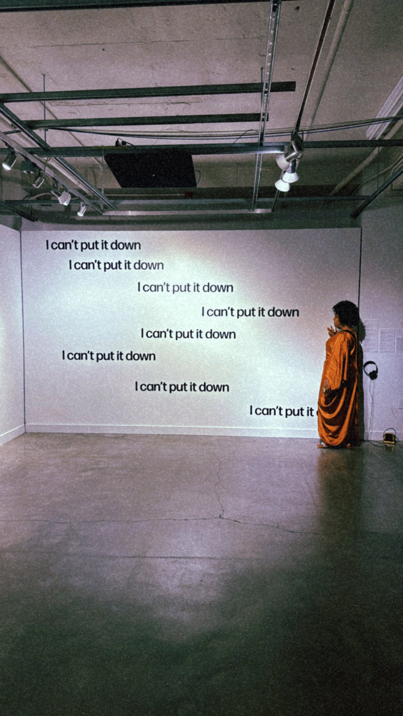 A Black woman with a curly Afro and wearing a in a long gold dress looks off camera at a white wall with the words, ‘I can’t put it down’ in black vinyl.