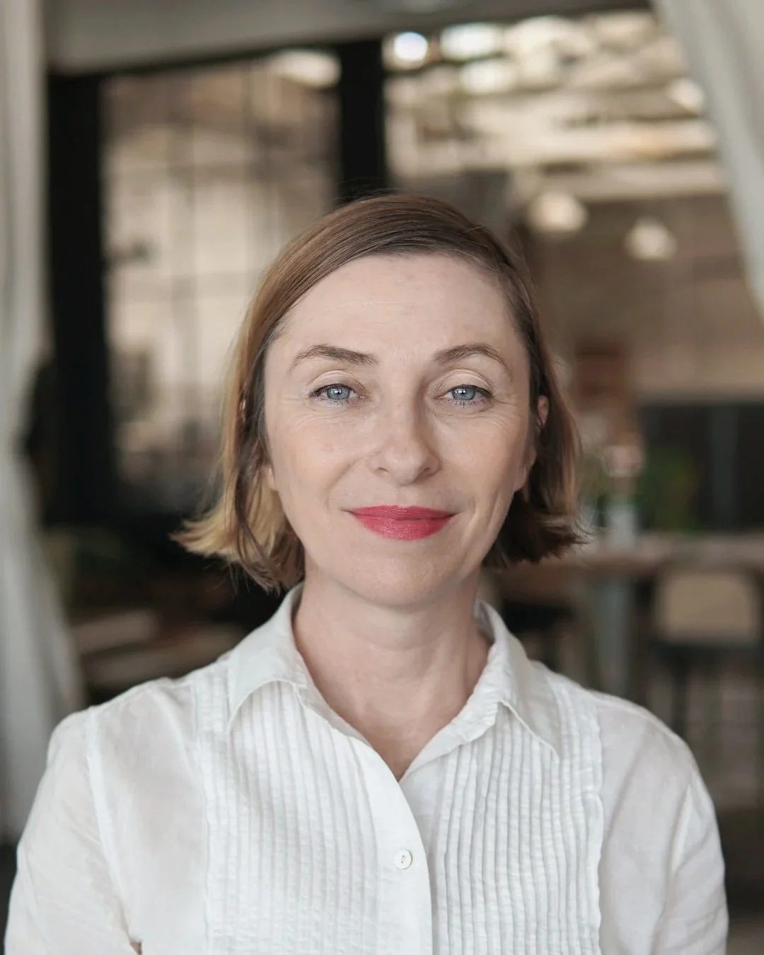 Close-up of a woman with short brown hair, blue eyes, and red lipstick, wearing a white collared shirt, smiling softly, in an indoor setting with a blurred background.