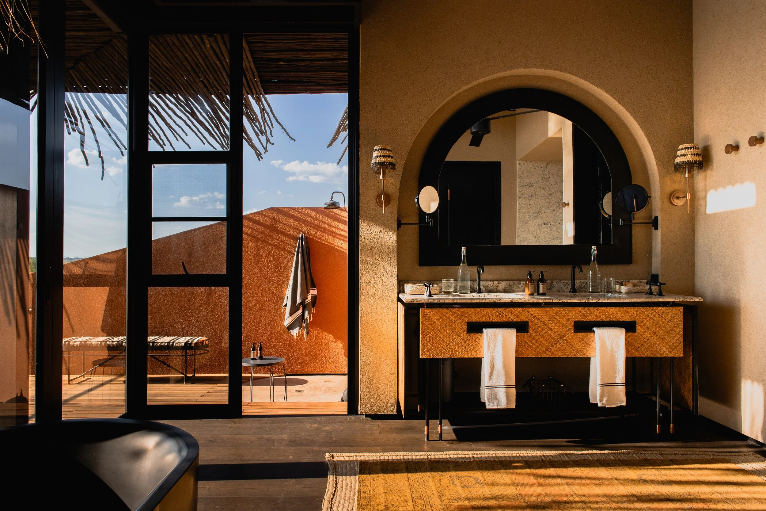 Interior view of a bathroom with a large mirror, dark frame, and beige textured walls, illuminated by two wall-mounted lamps with striped lampshades. Through an open door, a balcony or outdoor area is visible, featuring a terracotta-colored wall, a s