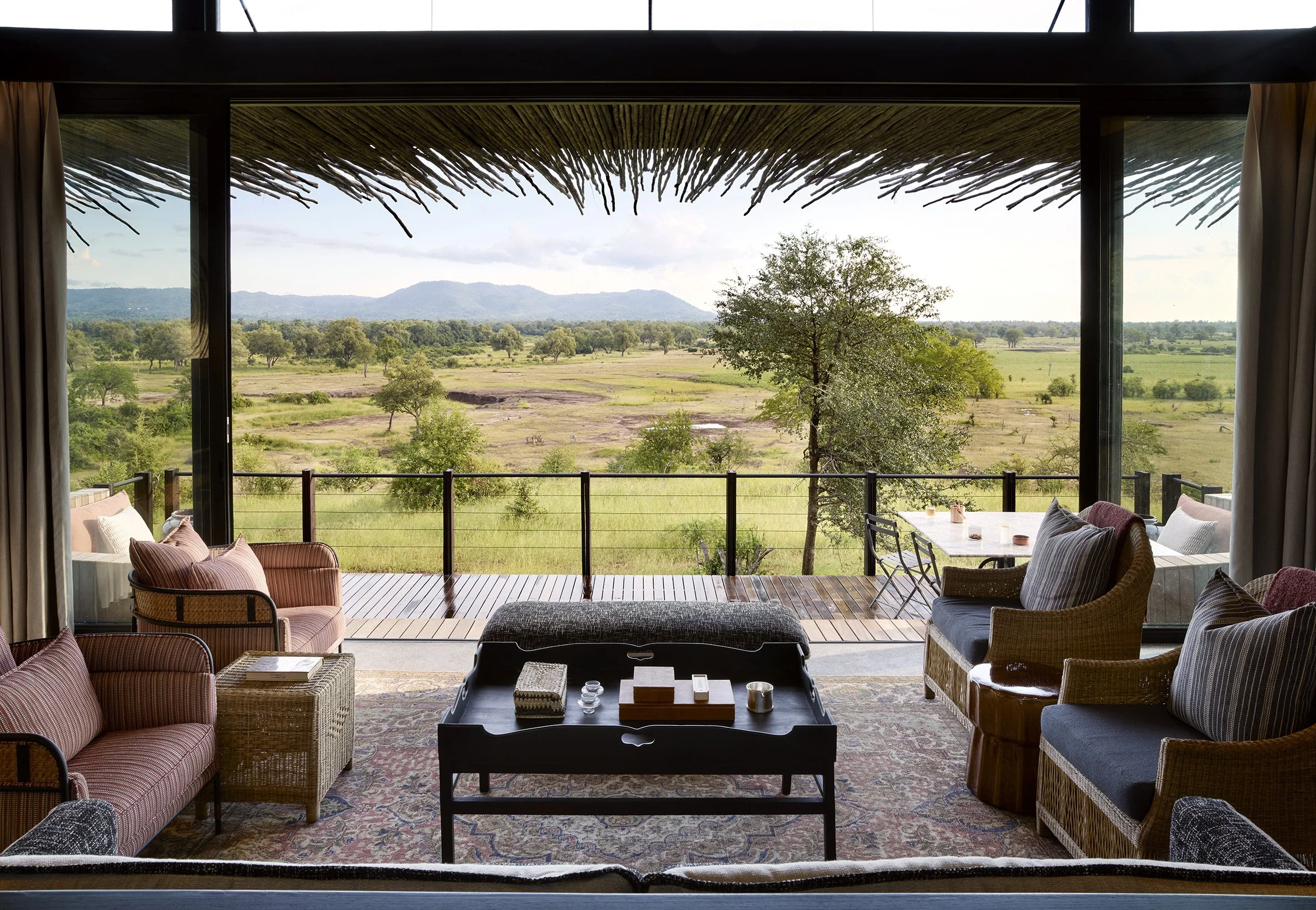 View of a landscape with trees, grass, and hills through large open glass doors from a room interior. Inside, there are sofas and chairs around a coffee table on a patterned rug.